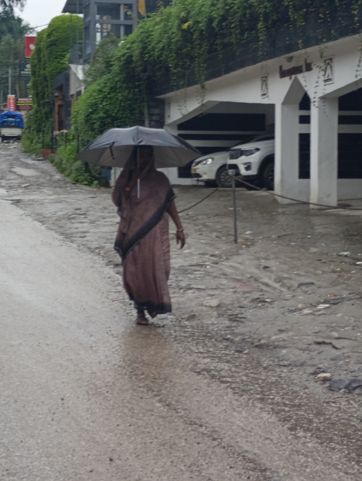 A person walks alone with an umbrella on a wet road in Aleo, India. The person, wearing a brown garment and a dark scarf, is covered by a black umbrella, suggesting it is raining. They are walking on a paved road that is wet and reflects the overcast sky. In the background, there is a white building with arched openings and parked cars. Lush green vegetation covers the side of a building behind the person, and a small vehicle is visible further down the road. The overall environment appears to be an urban or semi-urban area experiencing rain.