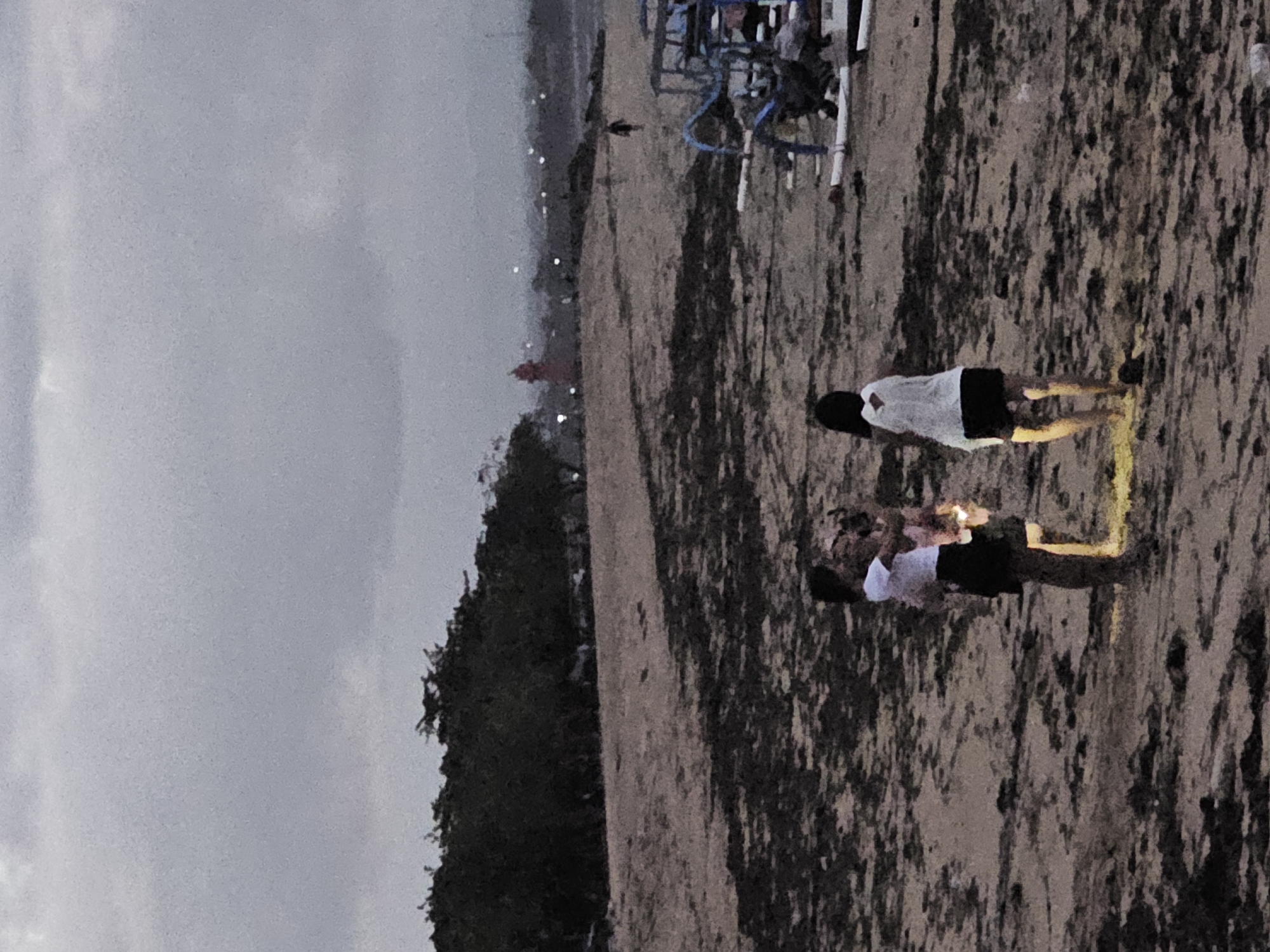 Two young females stand on a sandy beach in Denpasar, Indonesia, at dusk. One is holding a smaller child and both appear to be looking at something in front of them. The other female stands with her back to the camera, facing the water. The beach is covered in dark debris, likely seaweed. In the distance, a red tower is visible, along with a treeline and the lights of the city. Several fishing boats are pulled up onto the sand on the right side of the image. The sky is overcast.