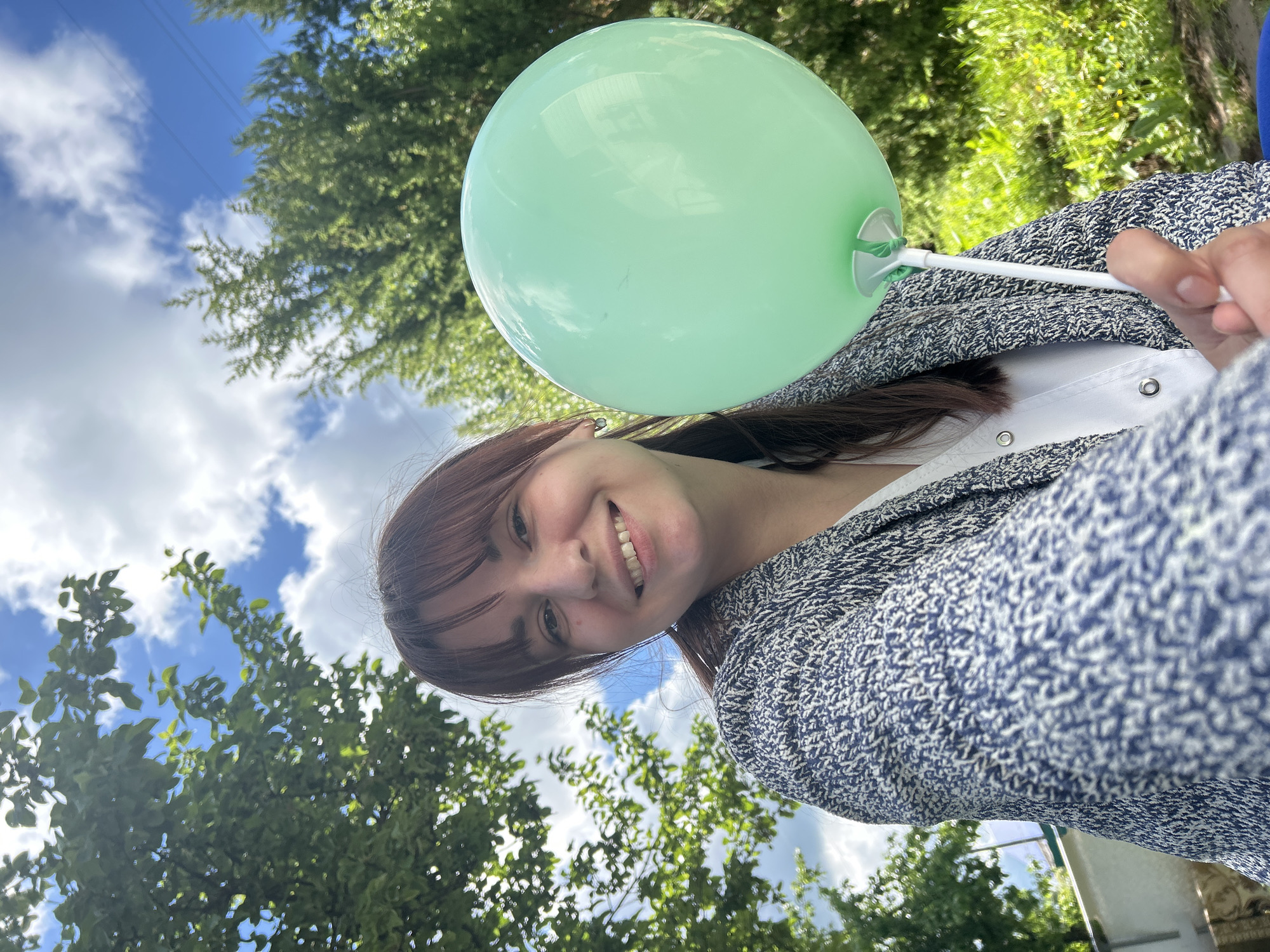 A woman with dark hair and bangs smiles, holding a pale green balloon on a white stick. She wears a white top and a blue patterned cardigan. The background features green trees and a blue sky with white clouds. A power line is visible in the sky. Location details cannot be reliably determined from the image.