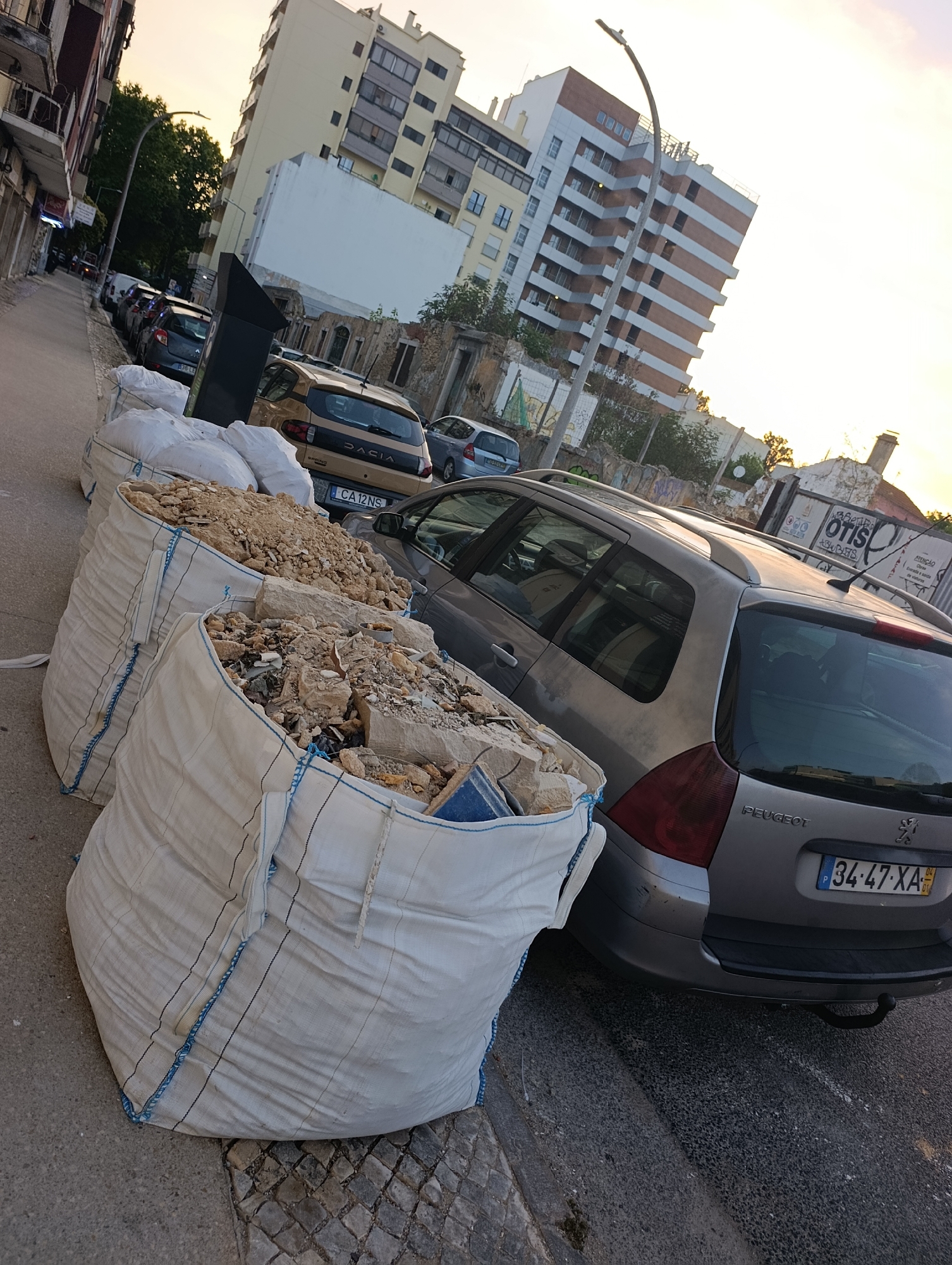 In Setúbal, Portugal, on a street with parked cars and buildings, construction debris is piled high in large white bags. The bags are situated on the sidewalk and partially obstruct the view of parked vehicles, including a silver Peugeot station wagon with license plate 34-47-XA. The scene appears to be in a residential or urban area, with several buildings visible in the background. The sky is bright, suggesting it is either sunrise or sunset.