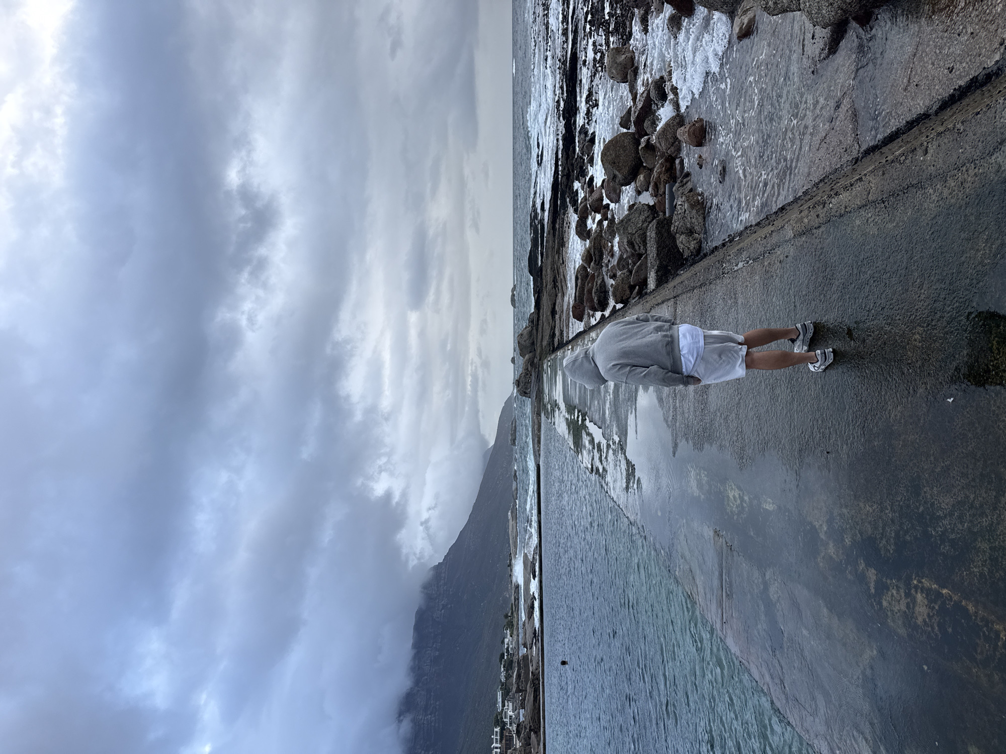 A person in a gray hoodie and white shorts stands on a concrete platform beside a tidal pool. The pool extends to the ocean's edge, with rocks and crashing waves. In the background, a mountain obscured by clouds is visible, with buildings at its base. The sky is overcast. This scene is located in Cape Town, South Africa.