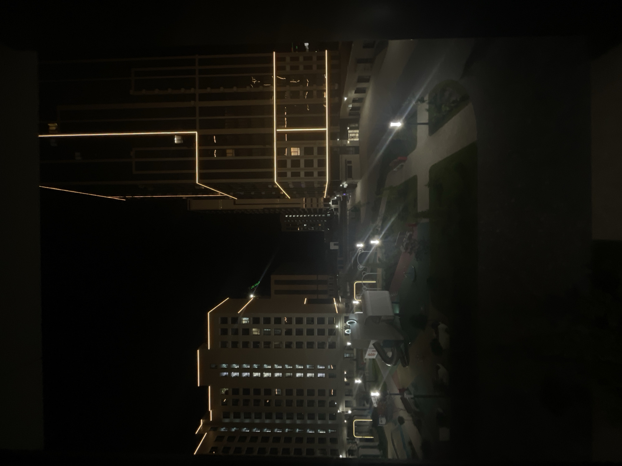 The image depicts a nighttime view of a modern residential complex in Stavropol, Russia. The architecture features tall, multi-story buildings with illuminated facades, highlighted by warm-toned LED strip lighting outlining the edges of the structures and balconies. In the foreground, a children's playground is visible with various play structures, including a slide, and surrounded by pathways and green spaces. Streetlights illuminate the courtyard, casting a glow on the paved areas and the playground. The scene is framed by dark borders, suggesting the photo was taken from a window.