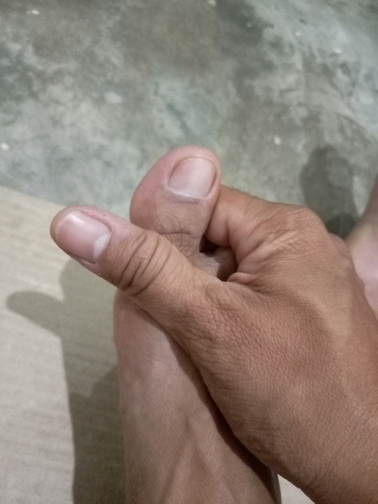 The image shows a close-up view of a person's foot and hand. The foot's toes are visible, with one toe being held by the thumb and index finger of the other hand. The skin appears to be light brown. The background is a textured, light gray surface, possibly concrete or a similar material. The lighting suggests an indoor environment, with shadows cast by the hand and foot on the surface. There are no specific landmarks or details in the image that would indicate a location in Batam, Indonesia.