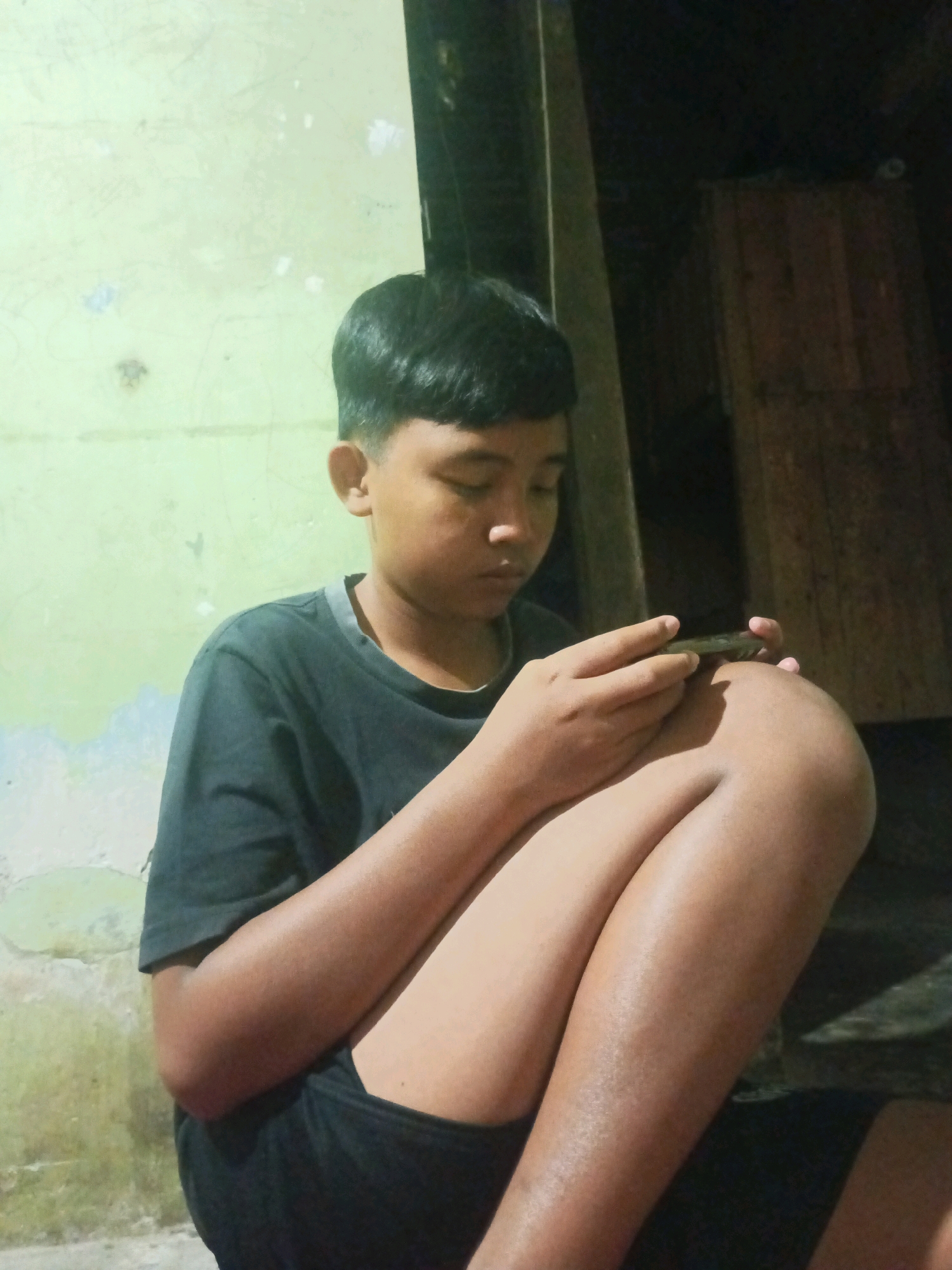 A young person, appearing to be of East Asian descent, with short black hair and medium skin tone, is sitting with their knees drawn up to their chest. They are wearing a dark t-shirt and dark shorts. The person is looking down and holding a dark-colored mobile phone in their hands, seemingly engaged with it. The background consists of a lightly textured, pale green wall on the left, with some darker patches. To the right, there is a wooden structure, possibly a door or a partition, and the area appears to be dimly lit, suggesting an indoor or covered outdoor setting. The overall impression is of a casual, everyday moment. The image was taken in Ngrendeng, Indonesia.