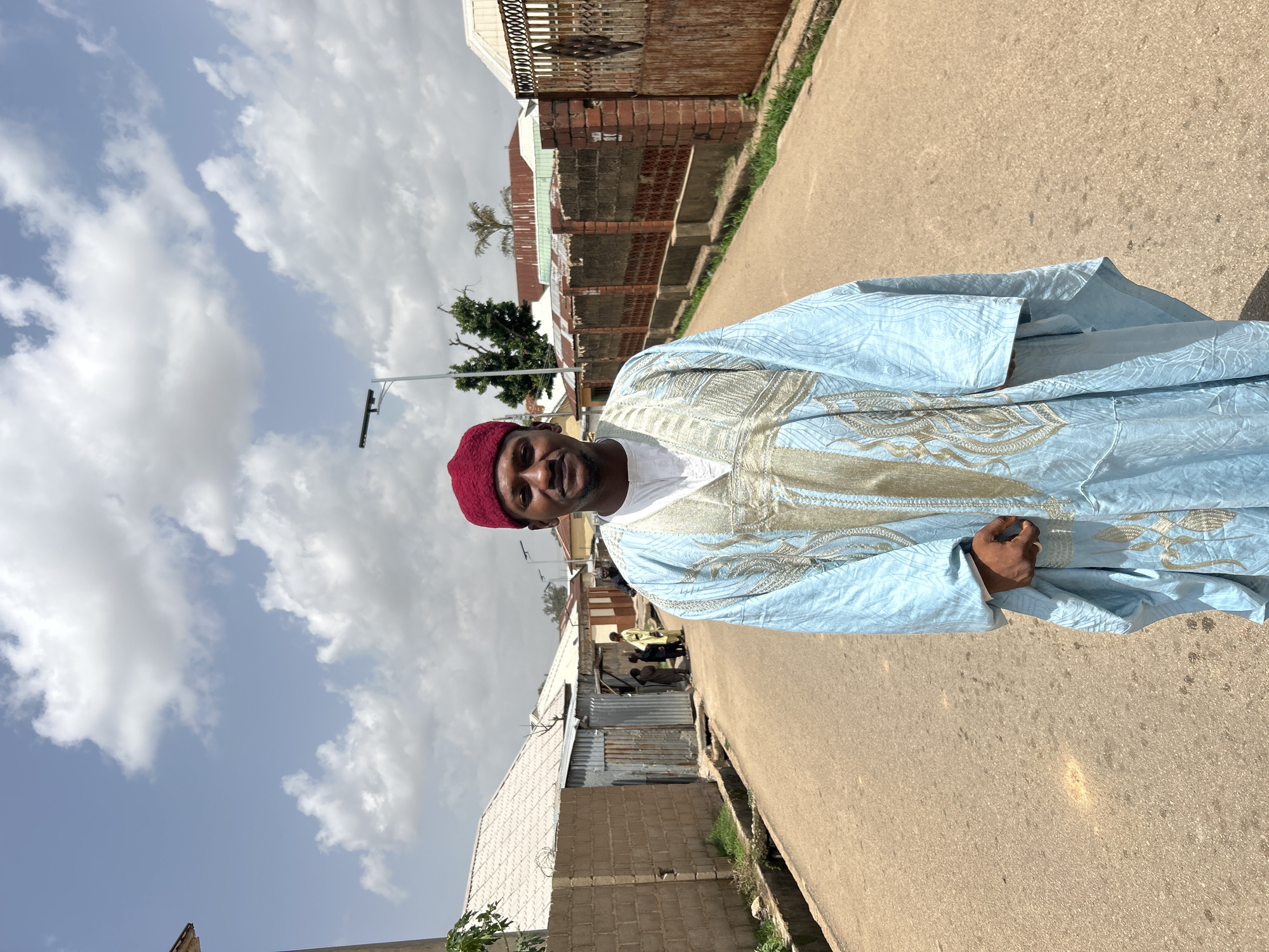 A man stands on a paved street in Jos, Nigeria. He wears a light blue traditional garment with gold embroidery and a maroon cap. Behind him, a mix of residential buildings is visible, some constructed from brick and others from corrugated metal. The sky is partly cloudy. Other people can be seen in the background, further down the street.