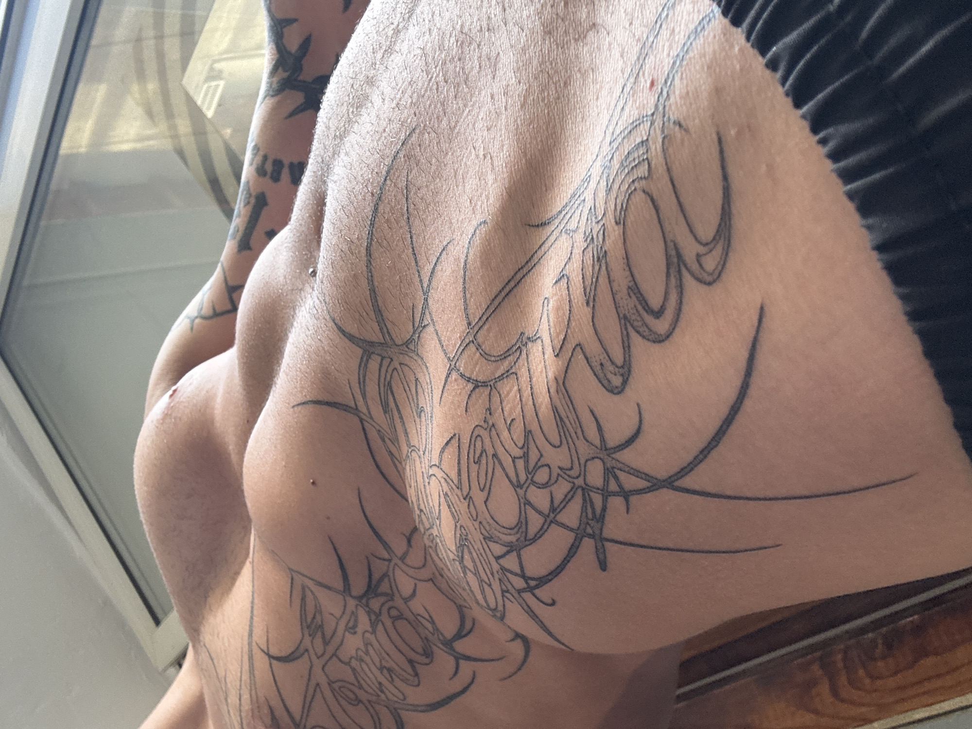 The image is a close-up, low-angle shot of a person's torso and arm, featuring extensive black tribal-style tattoos. The person is lying down, with their back muscles clearly defined. Their skin is light-toned and appears to have some light hair growth. A silver piercing is visible on their abdomen. The background shows a reflection in a window, suggesting an indoor setting with natural light. The bottom of the frame shows a glimpse of dark fabric, likely clothing. There is no discernible location in the city of Sofia, Bulgaria, present in the image.