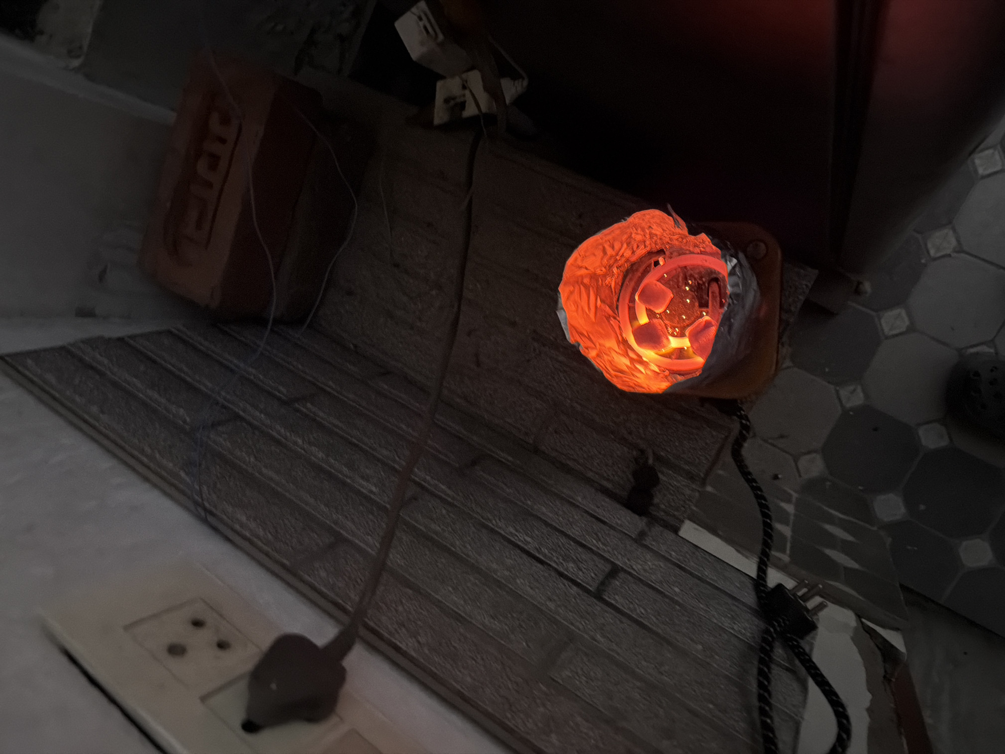 The image depicts a close-up, high-angle view of a heating element with glowing red coals, likely used for shisha or tobacco, placed inside a foil-lined container. The container sits on a patterned tiled floor. To the left of the heating element, an electrical plug is inserted into a white wall socket. The wall behind the socket has a textured, possibly wallpapered surface. Further to the right, a red brick with embossed lettering is visible, suggesting an urban setting. Wires are visible running across the scene, connecting the heating element to a power source and also near the brick. The overall lighting is dim, with the intense red glow of the coals being the primary light source in the immediate vicinity. The scene appears to be indoors, in a domestic or informal setting, possibly in Delhi, India, given the type of electrical socket and brick commonly found in the region.