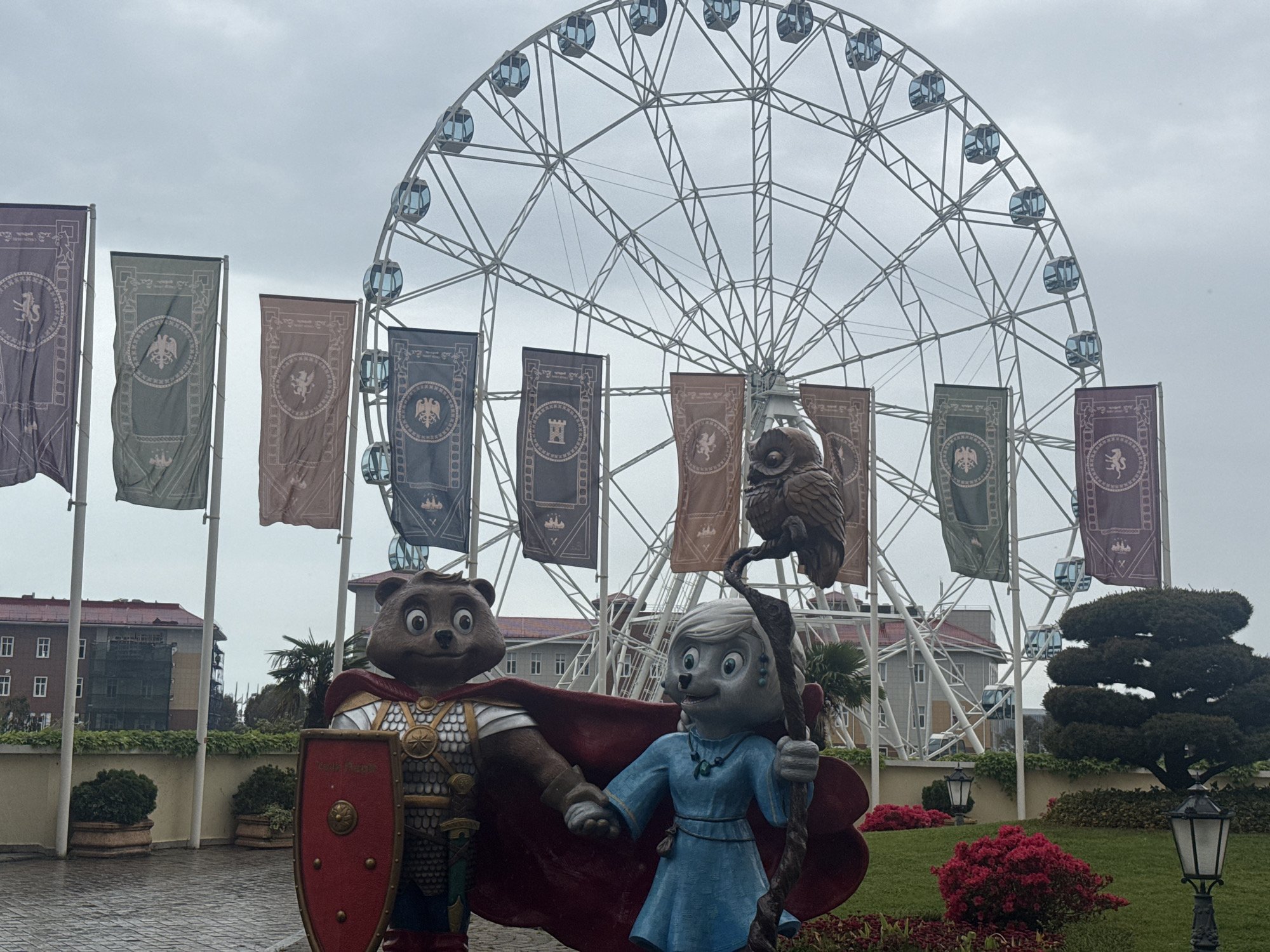 The image shows a scene in Sirius, Russia, featuring a large Ferris wheel in the background, likely a tourist attraction. In the foreground, there are statues of anthropomorphic animal characters dressed in medieval-style clothing. The male character, a bear, is wearing armor and holding a shield, while the female character, possibly a wolf or fox, is in a blue dress and holding a staff with an owl on top. Several banners with different symbols are positioned behind the statues and in front of the Ferris wheel. The setting appears to be a park or garden, with well-maintained greenery, flowerbeds, and trimmed bushes. The sky is overcast, suggesting a cloudy day. Buildings can be seen in the background behind the Ferris wheel and to the left of the scene.
