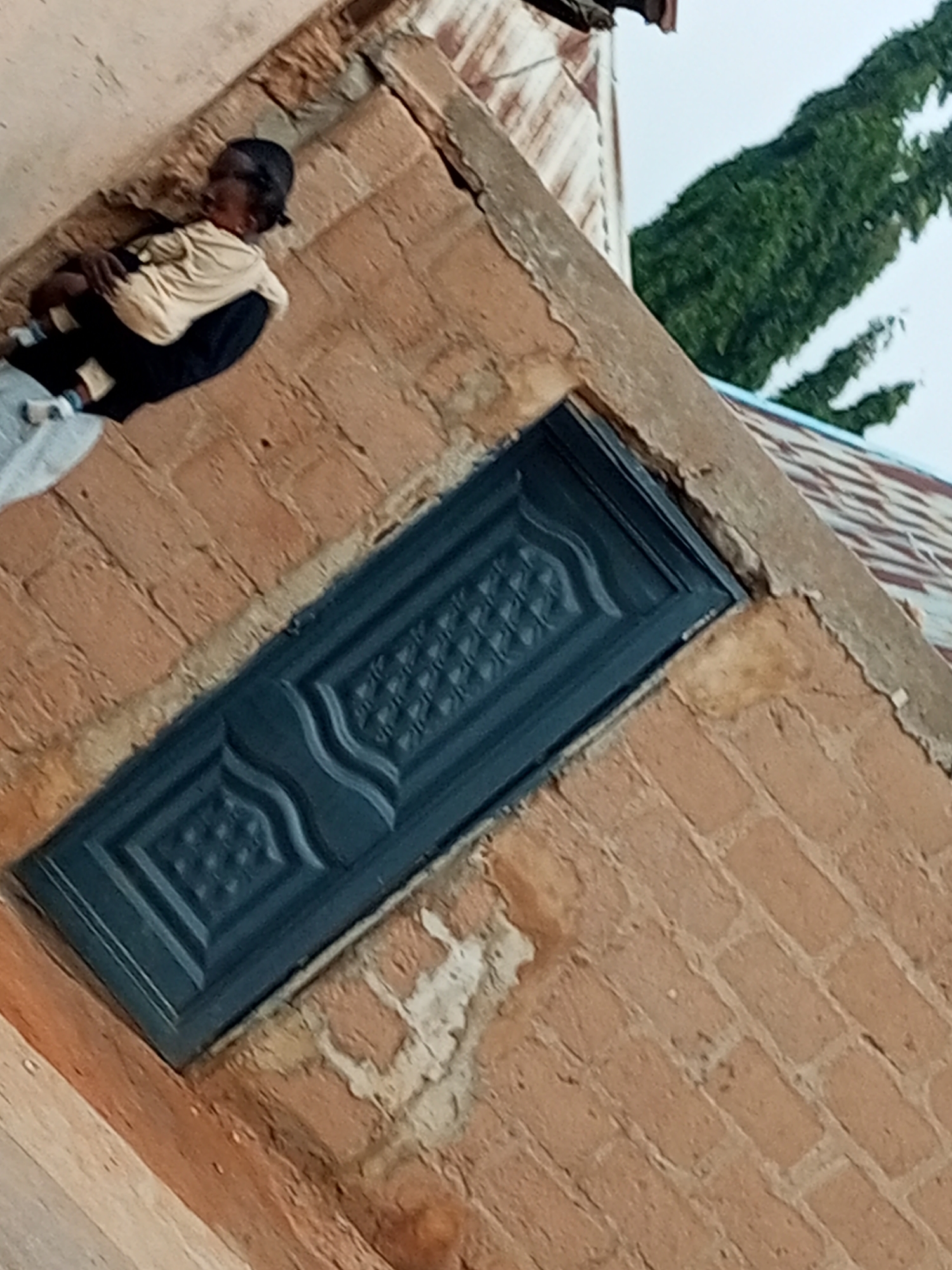 In Tumfure, Nigeria, a child is depicted lying down on a brick surface. The child is wearing a light-colored shirt over a dark garment and a dark cap. The child appears to be sleeping or resting. Behind the child, a dark-colored metal door with an ornate pattern is visible, set into a brick wall. The environment suggests an outdoor setting, possibly a yard or an alleyway, with hints of a tiled roof and foliage in the background.