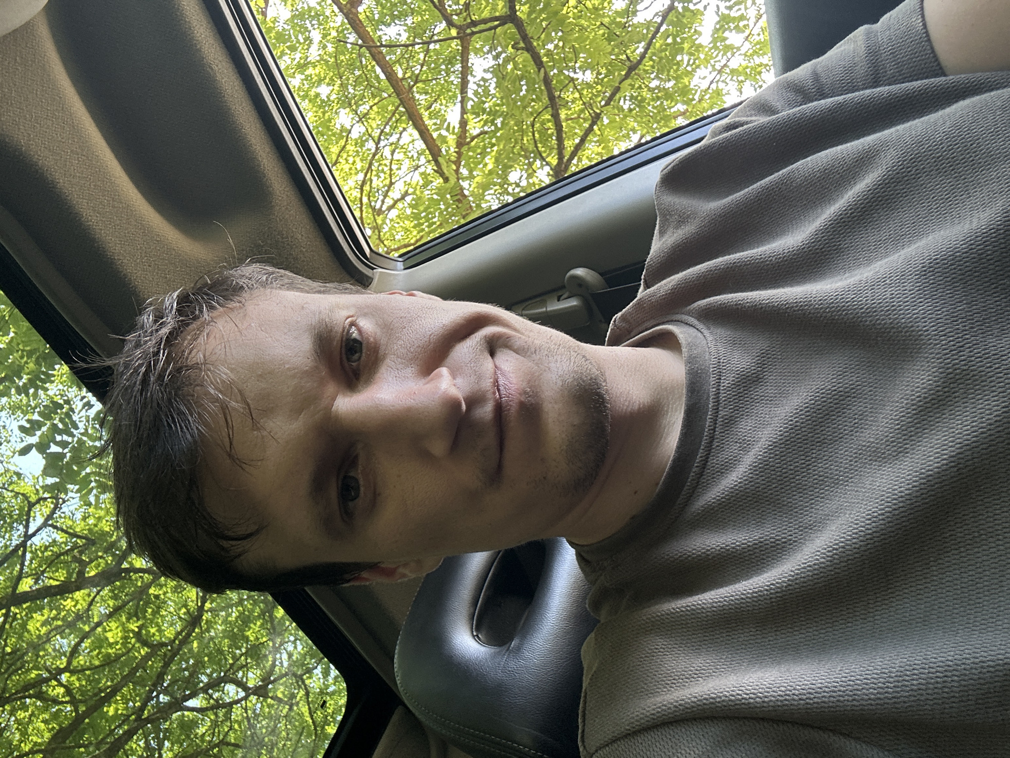In Makiv, Ukraine, a light-skinned male with short brown hair and stubble is pictured from the chest up, seated in a car. He is wearing a textured brown t-shirt. The interior of the car is visible, including a dark gray headrest and the car's ceiling. Sunlight filters through green foliage and branches of trees visible through the car's open sunroof and window. The man is looking directly at the camera with a slight smile.