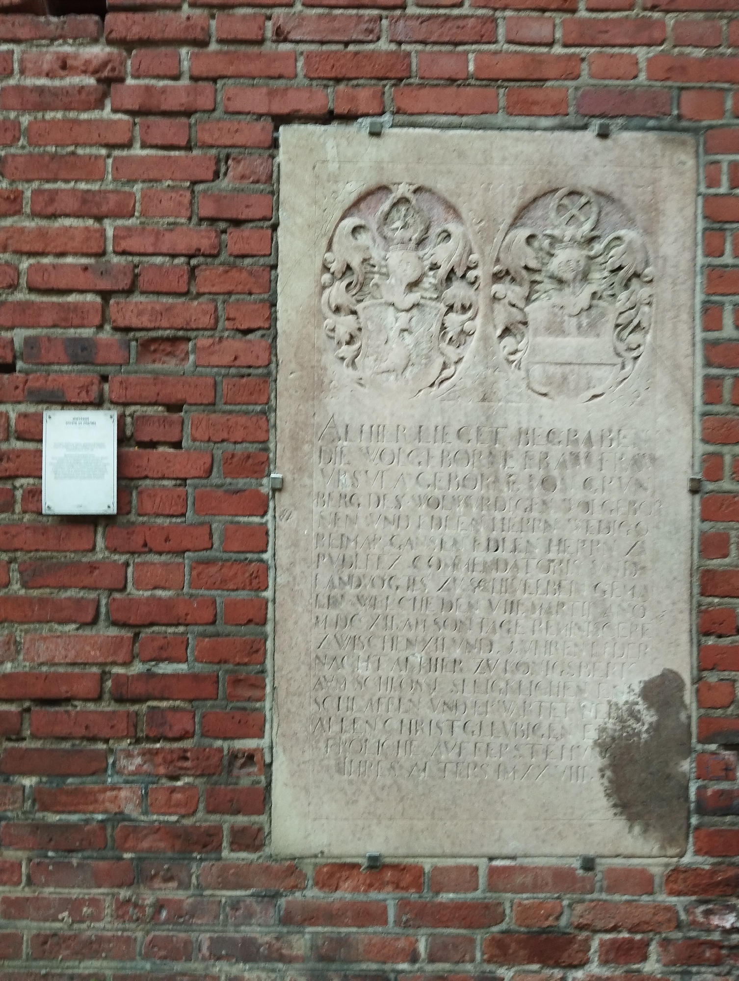 The image shows a close-up of a red brick wall with a large stone tablet embedded in it. The tablet is inscribed with text and features two crests at the top, each enclosed in a circular, decorative frame. The text on the tablet appears to be in German, likely an epitaph or historical inscription. To the left of the stone tablet, a smaller white rectangular plaque is attached to the brick wall. The brickwork is weathered, suggesting age. The scene is set outdoors, with the natural lighting casting shadows on the stone tablet. The location is the city of Kaliningrad, Russia, specifically on the building of the former Königsberg Castle.