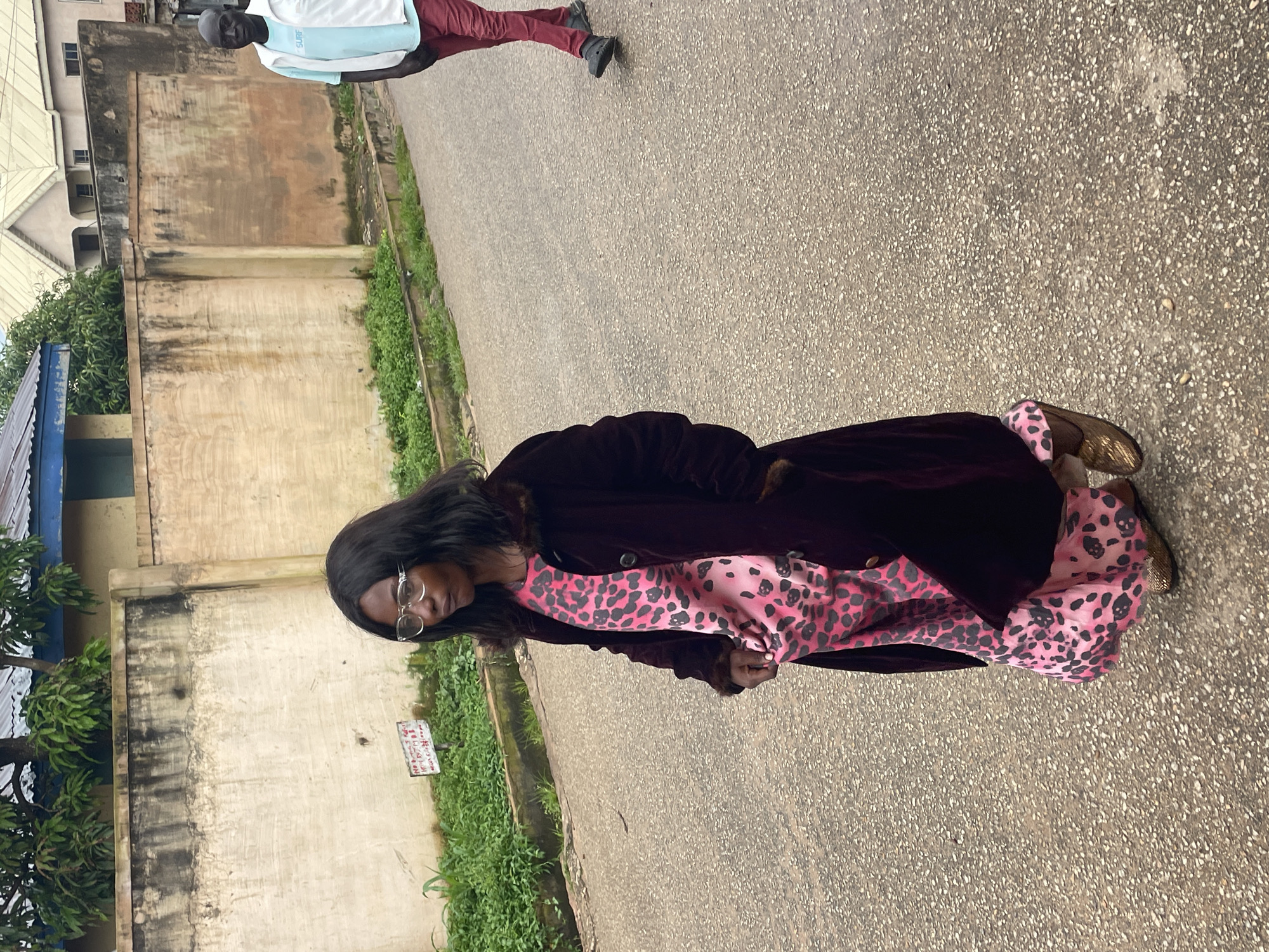 In Jos, Nigeria, a medium shot captures two individuals walking on a paved street. In the foreground, a woman with dark, shoulder-length hair wears glasses, a dark maroon coat with fur trim, and a pink dress with a black leopard print. She is looking to her left. In the background, to the right of the woman, a man with dark skin is walking away from the camera. He is wearing a light blue short-sleeved shirt with the word "SURF" visible on it and dark red pants. Behind the figures, a beige concrete wall runs horizontally across the frame, with green vegetation growing along its base. A building with a blue-tiled roof is partially visible above the wall on the left, and other buildings are seen in the distance on the right. The street is asphalt, with visible gravel and some reflective patches, suggesting it may be damp. A faded red and white sign is attached to the wall on the left.