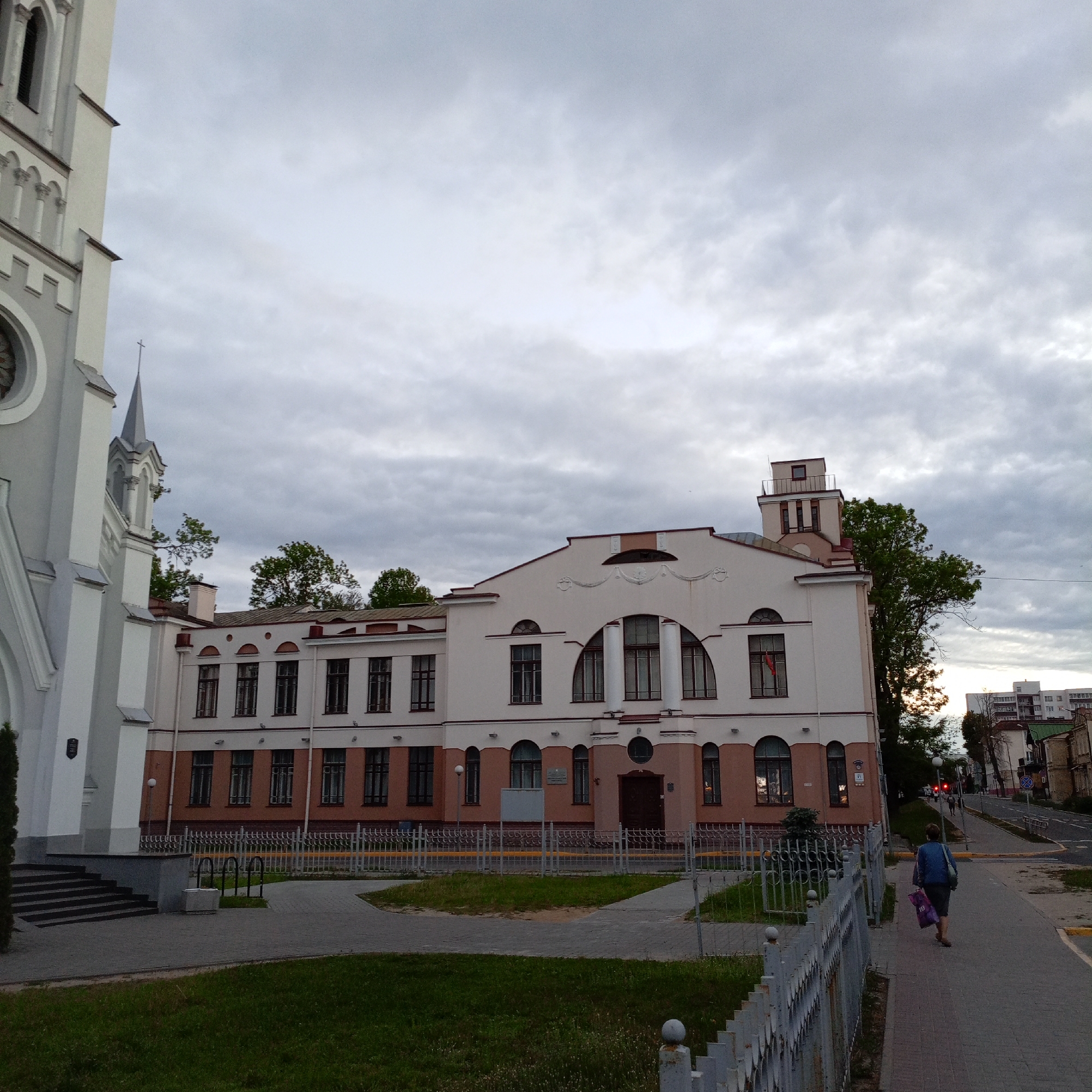 The photograph captures a street scene in an urban area under a cloudy sky. A woman wearing a blue jacket and carrying a purple bag walks along a sidewalk adjacent to a street. A white fence runs parallel to the sidewalk. To the left, a portion of a white church with gray trim and steps is visible. Adjacent to the church is a two-story building with a distinctive architectural style featuring arched windows and a small tower-like structure on its roof. A small patch of green lawn is visible between the sidewalk and the building. In the background, other buildings and a street with a crosswalk and a traffic light are seen.