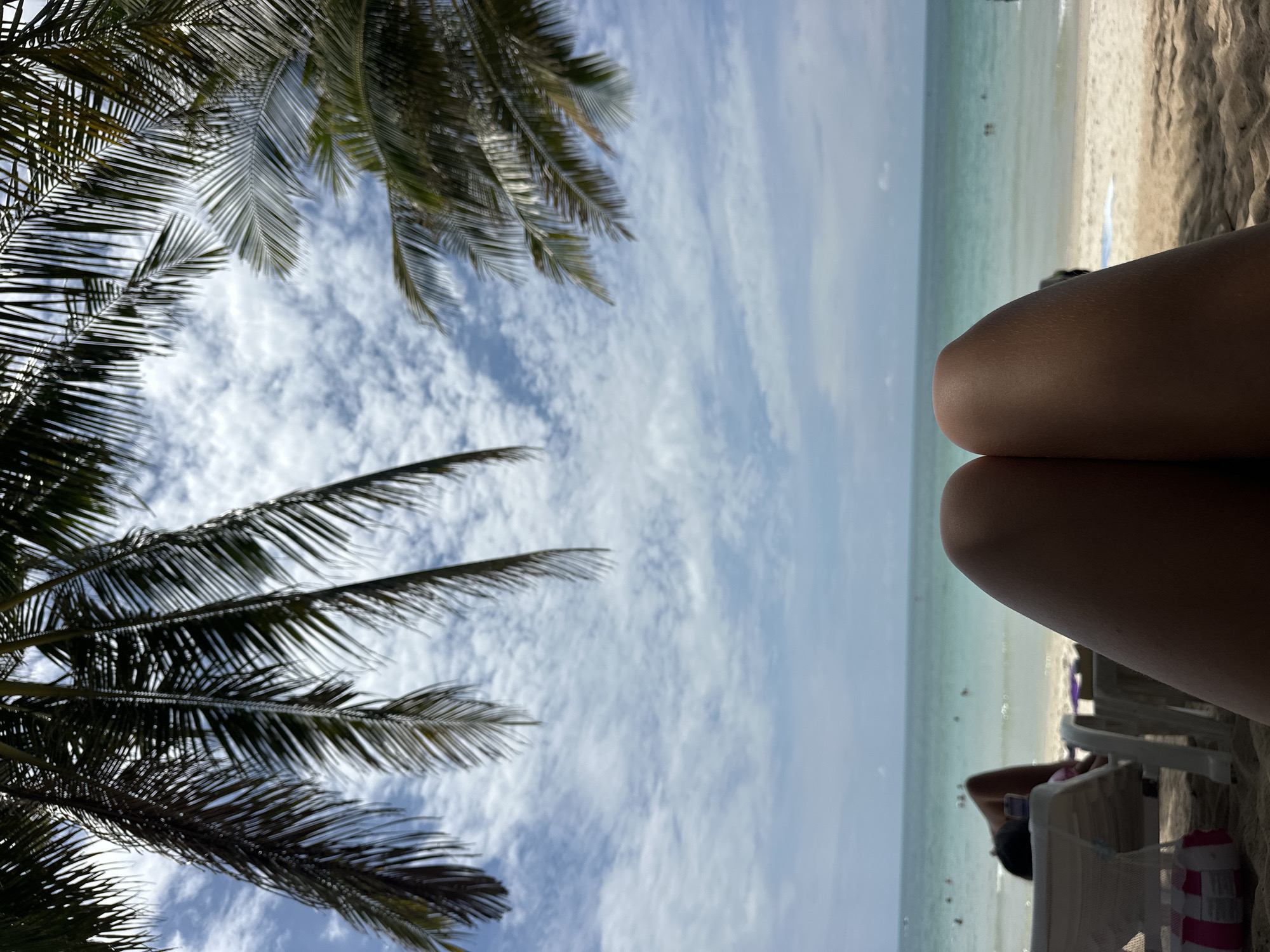 The image shows a first-person perspective of someone relaxing on a beach in Koh Samui District, Thailand. In the foreground, a person's bare legs are visible, suggesting they are lying down. Above the legs, palm fronds frame a view of a bright blue sky filled with scattered white clouds. Beyond the beach, a turquoise ocean stretches to the horizon. Several people can be seen in the water and on the sand in the background, along with lounge chairs. The sand is light in color. The overall scene depicts a serene beach setting on a sunny day.