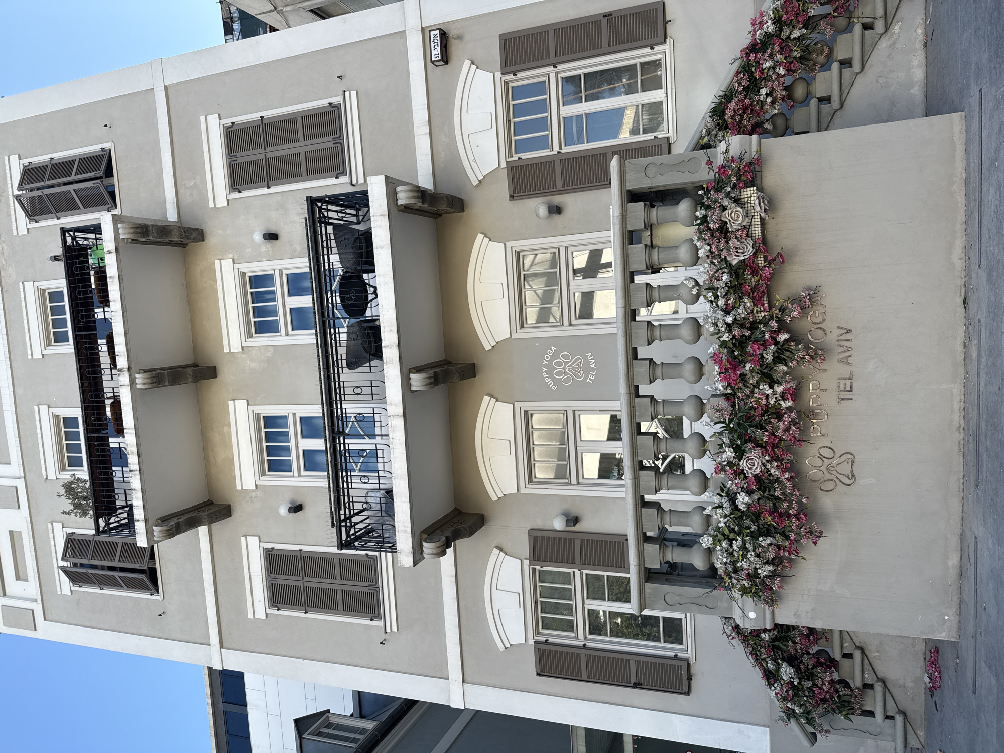 The image shows the exterior of a light gray building with white trim in Tel Aviv, Israel. The building has multiple floors with several windows, many featuring dark brown shutters that are partially or fully closed. Balconies with black metal railings are present on the upper floors. The ground level features a concrete facade with decorative elements. Two staircases, adorned with abundant floral arrangements of pink and white flowers, lead up to the entrance. Above the stairs, a sign with a paw print logo and the text "PUPPY YOGA TEL AVIV" is visible. A street sign with Hebrew text and the number "52" is mounted on the wall. The sky is clear and blue. No people are visible in the image.