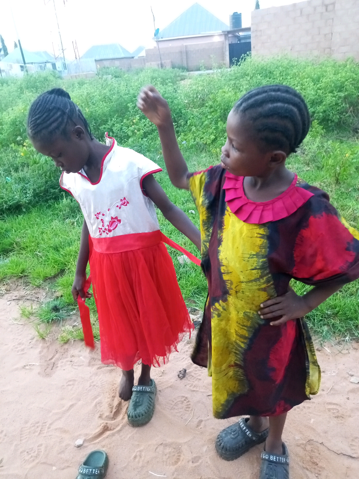 Two young girls, appearing to be of African descent, are standing on a sandy surface next to a grassy area. The girl on the left is wearing a white dress with red trim and floral embroidery, and a red skirt. She is holding a red ribbon. The girl on the right is wearing a colorful dress with a yellow, red, and black tie-dye pattern and a ruffled pink neckline. She has her hand on her hip. Both girls are wearing dark-colored clogs. In the background, there are buildings and vegetation, suggesting an outdoor setting in a residential area. The scene is lit by natural daylight. The presence of buildings and vegetation is consistent with a location in Tempure, Nigeria.