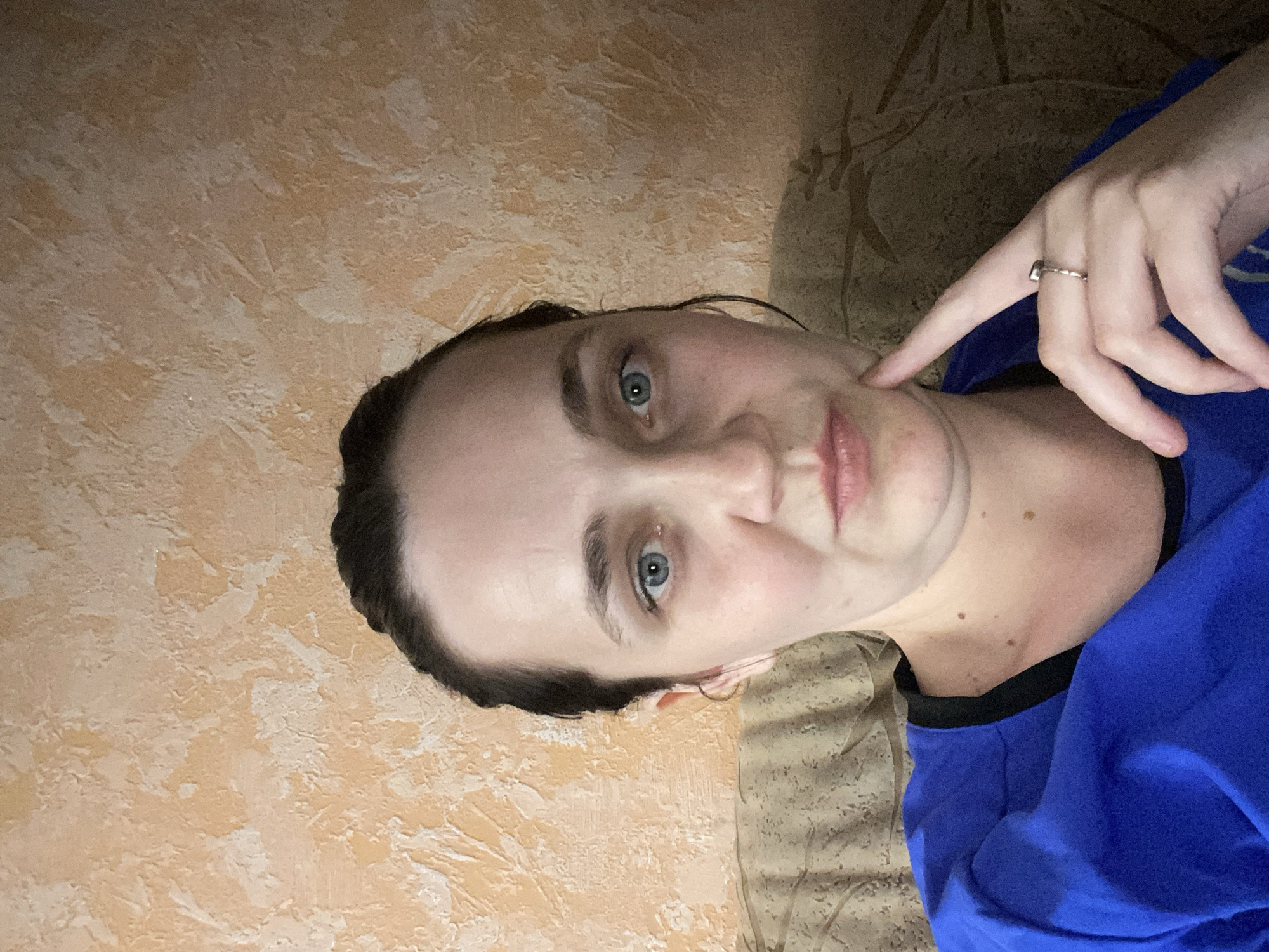 An indoor, eye-level, medium shot shows a woman looking directly at the camera. The woman has light skin, dark brown hair pulled back into a bun, and blue eyes. She is wearing a royal blue shirt with a black trim around the neckline. Her right hand is resting on her chin, and she is wearing a silver ring on her right ring finger. Behind her, the wall has an orange textured finish, and to her left, a portion of a brown patterned couch is visible. The lighting in the scene is soft, casting shadows on her face and the wall. The image is slightly blurry in some areas.