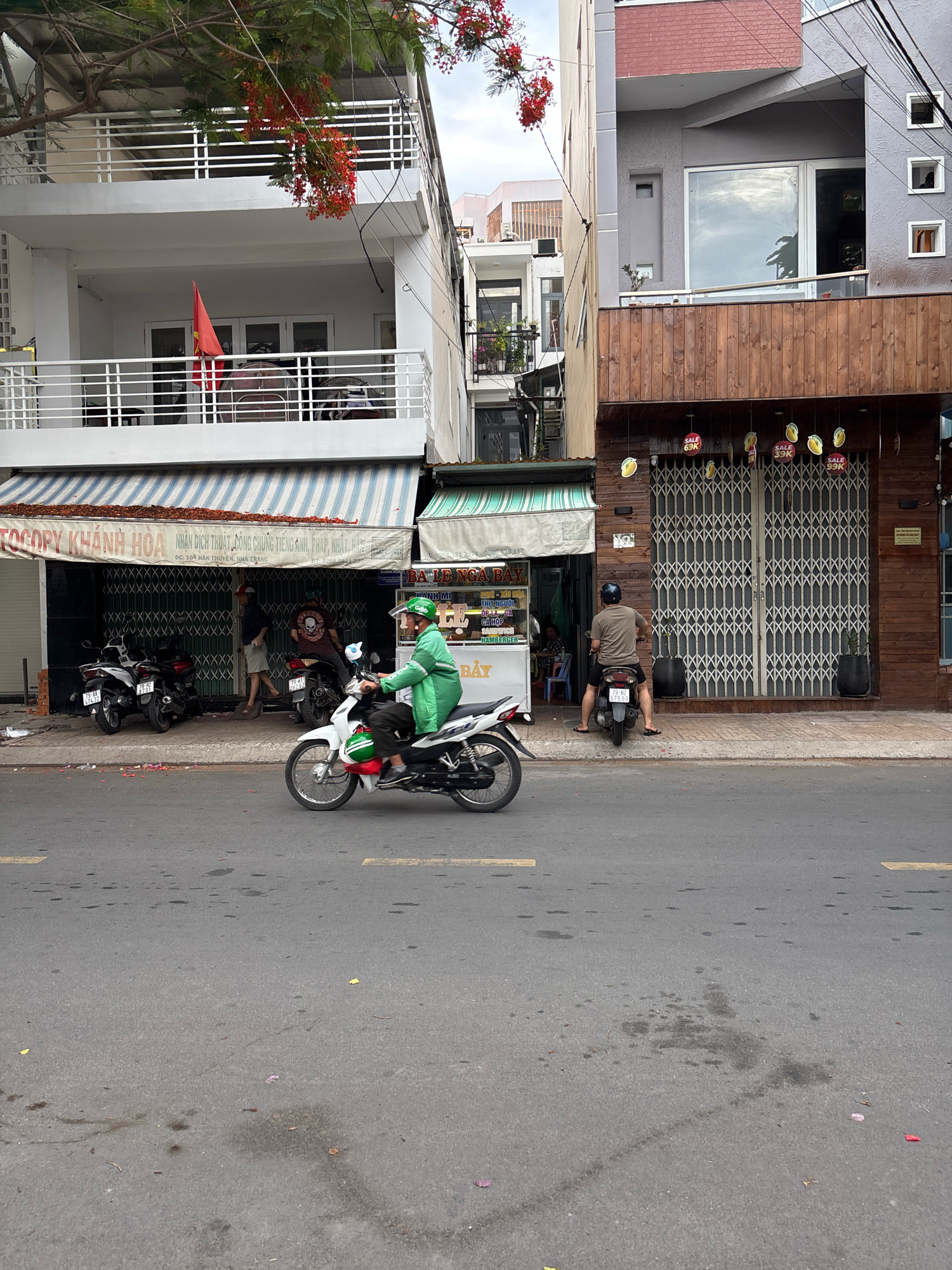 In Nha Trang, Vietnam, a street scene unfolds with a focus on a sidewalk and road. On the sidewalk, several motorcycles are parked. One white motorcycle, with a green-clad individual wearing a helmet, is in motion, heading towards the viewer. Another man on a dark scooter faces away from the viewer. Behind them, a building with a wooden facade features large, corrugated metal shutters. Several round signs with yellow fruit motifs and prices like "SALE 69K" are displayed above the shutters. To the left, a white building with balconies and a red Vietnamese flag is visible, with a striped awning over a storefront displaying text in Vietnamese. The street in the foreground is asphalt, showing some stains and tire marks. Red and pink petals are scattered on the sidewalk and street.