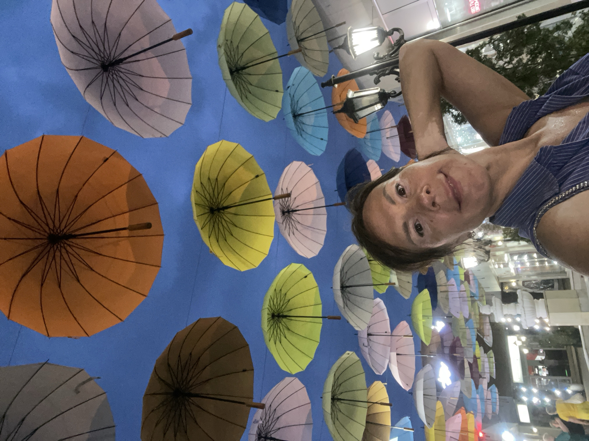 The image shows a woman taking a selfie outdoors at night, with a sky full of colorful umbrellas above her. The umbrellas are of various colors, including orange, yellow, green, pink, white, and blue, and are suspended in the air, creating a canopy. The woman is wearing a blue and white striped shirt and has her arm raised behind her head. In the background, there are buildings with illuminated windows and streetlights, suggesting an urban setting. A fountain and some people are visible in the lower part of the image. The scene appears to be located in the city of Sochi, Russia, specifically on a pedestrian street that has been decorated with umbrellas.