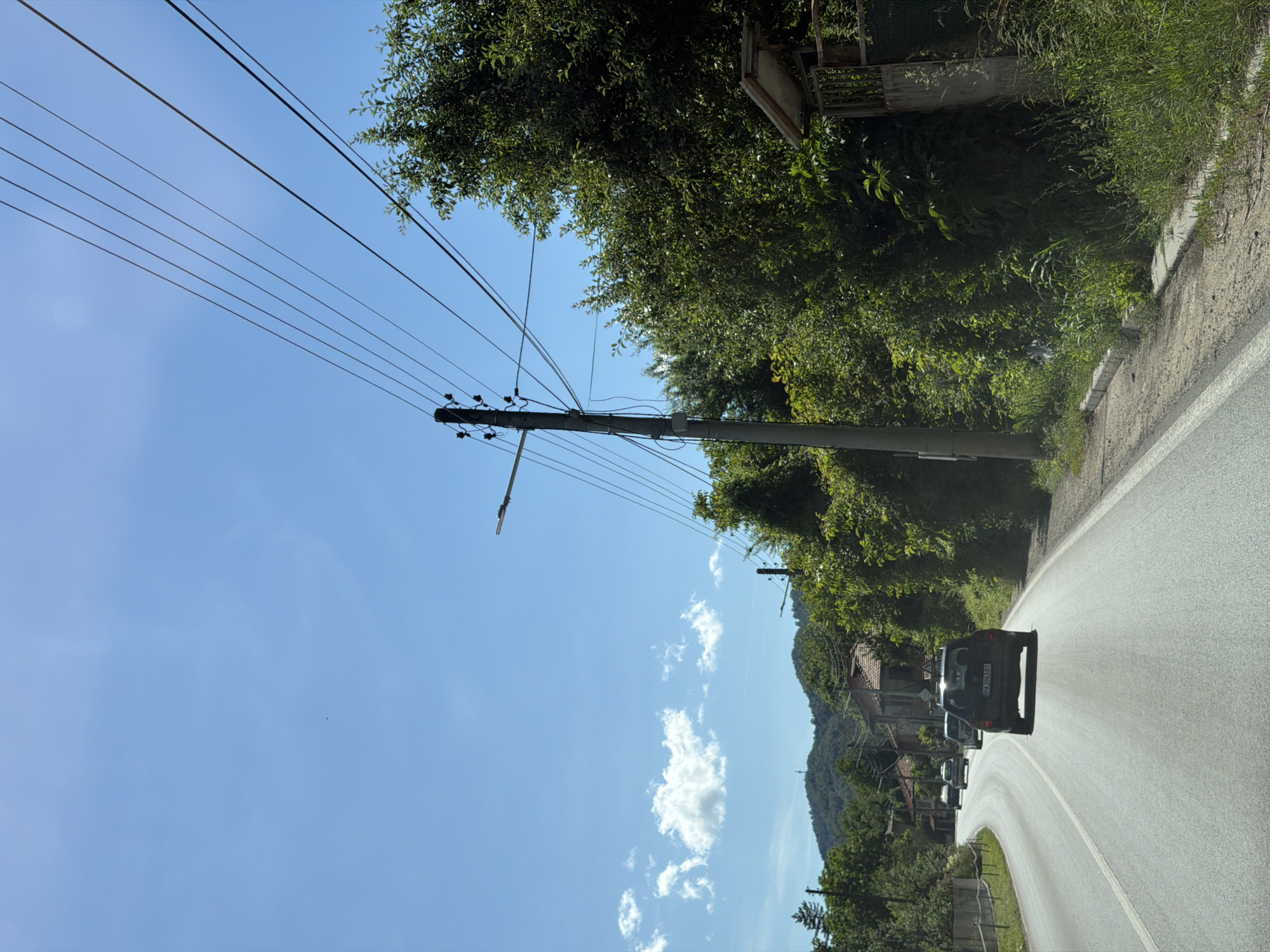The image depicts a rural road in Beli Osam, Bulgaria, under a clear blue sky with sparse clouds. Several cars are visible driving on the road, with one black SUV in the foreground. Overhead, power lines stretch across the frame, supported by a concrete utility pole. Dense green trees line the right side of the road, with some buildings and rolling hills in the background. The scene appears to be a sunny day, likely in a semi-urban or village setting.