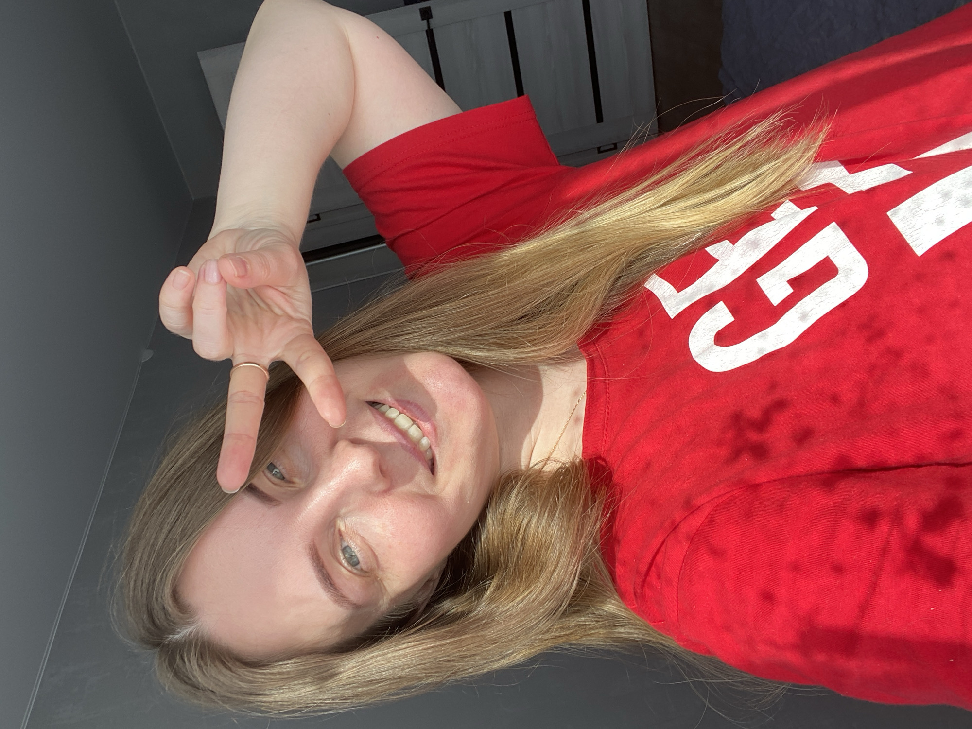 This image captures a female individual indoors in the city of Noviy, Russia. The subject has light blonde hair, fair skin, and blue eyes, and is smiling with teeth visible. She is wearing a red short-sleeved t-shirt featuring large white lettering, partially displaying "GE". A thin gold-colored necklace is visible around her neck. Her right hand is raised to her face, displaying a V-sign with the index and middle fingers, partially obscuring her face. A gold-colored ring is present on the ring finger of her right hand. Direct sunlight illuminates the subject from the right, creating sharp highlights and shadows. The background consists of a plain dark grey wall on the left. To the right, a dark-colored interior door with multiple horizontal light-colored panels is partially visible. The floor in the lower right is dark and indistinct.