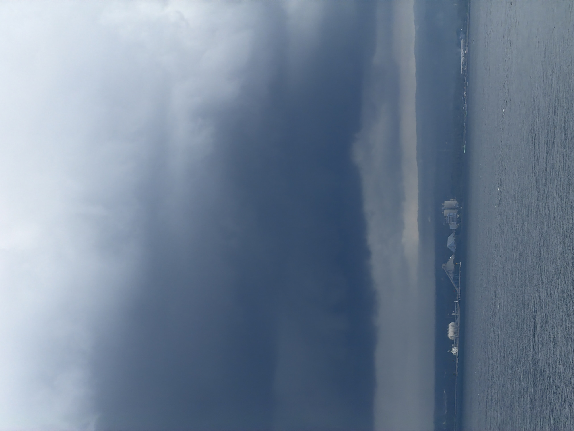 The image depicts a body of water, likely the sea, stretching horizontally across the lower portion of the frame. Along the shoreline, discernible structures suggest a port facility, possibly located in Gilimanuk, Indonesia. Dominating the left side of the image is a large, dark storm cloud. The sky above the sea is overcast.