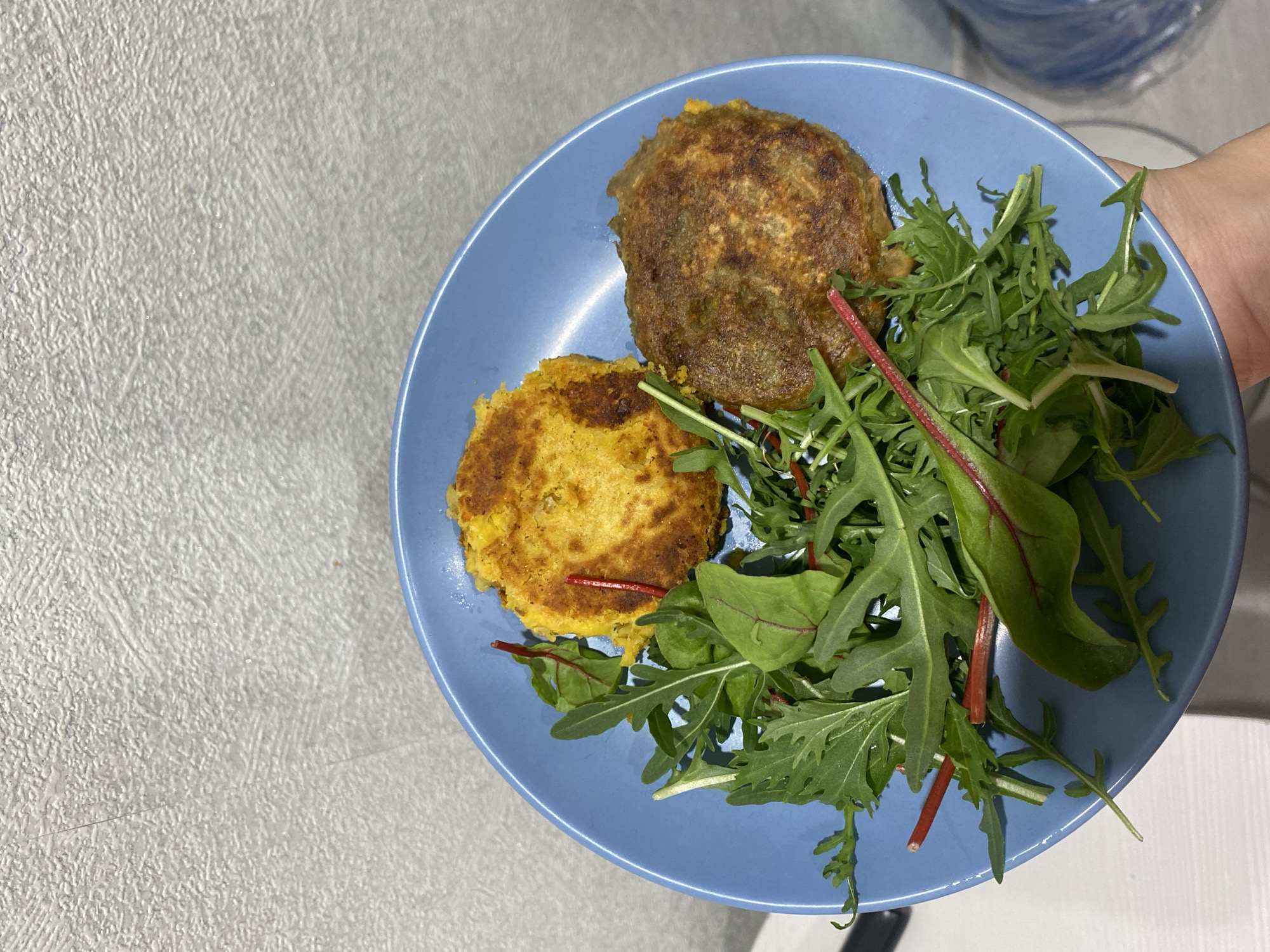 The image captures a close-up, overhead view of a meal served on a round, light blue ceramic plate. On the plate are two circular, pan-fried or baked patties. The patty in the lower-left is golden-yellow with a slightly coarse texture, while the one in the upper-right is darker, with a mottled brown and greenish-brown surface. Alongside the patties is a fresh mixed green salad, predominantly consisting of dark green, jagged-edged leaves, likely arugula, with some red-stemmed leaves integrated within, possibly chard or beet greens.
A hand, partially visible on the right side of the frame, is holding the plate, indicating a human presence presenting the meal. The plate is set against a light grey or off-white textured surface, likely a table or countertop. The lighting appears bright and natural, suggesting an indoor setting during daylight hours or a well-lit room. There is no visible text. The scene depicts a simple, prepared meal, captured in Chaikovsky, Russia.
