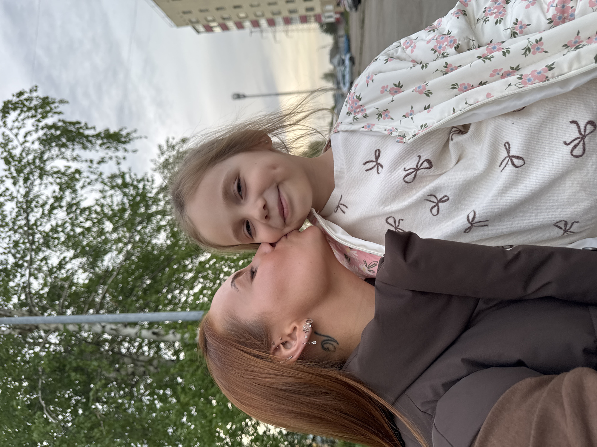 A photograph captures a moment of intimacy between an adult woman and a young girl, set against the backdrop of Chaikovsky, Russia. The woman, with auburn hair and a brown jacket, is positioned lower in the frame and appears to be kissing the girl's nose. The girl, with light blonde hair and a dimple, wears a white jacket with pink floral patterns and a white sweatshirt with brown bows. Behind them, there is a green tree and a gray pole, suggesting a park or public area. The background features a multi-story building with windows and parked cars, typical of an urban setting in Chaikovsky. The image is slightly tilted, giving a casual, snapshot aesthetic.