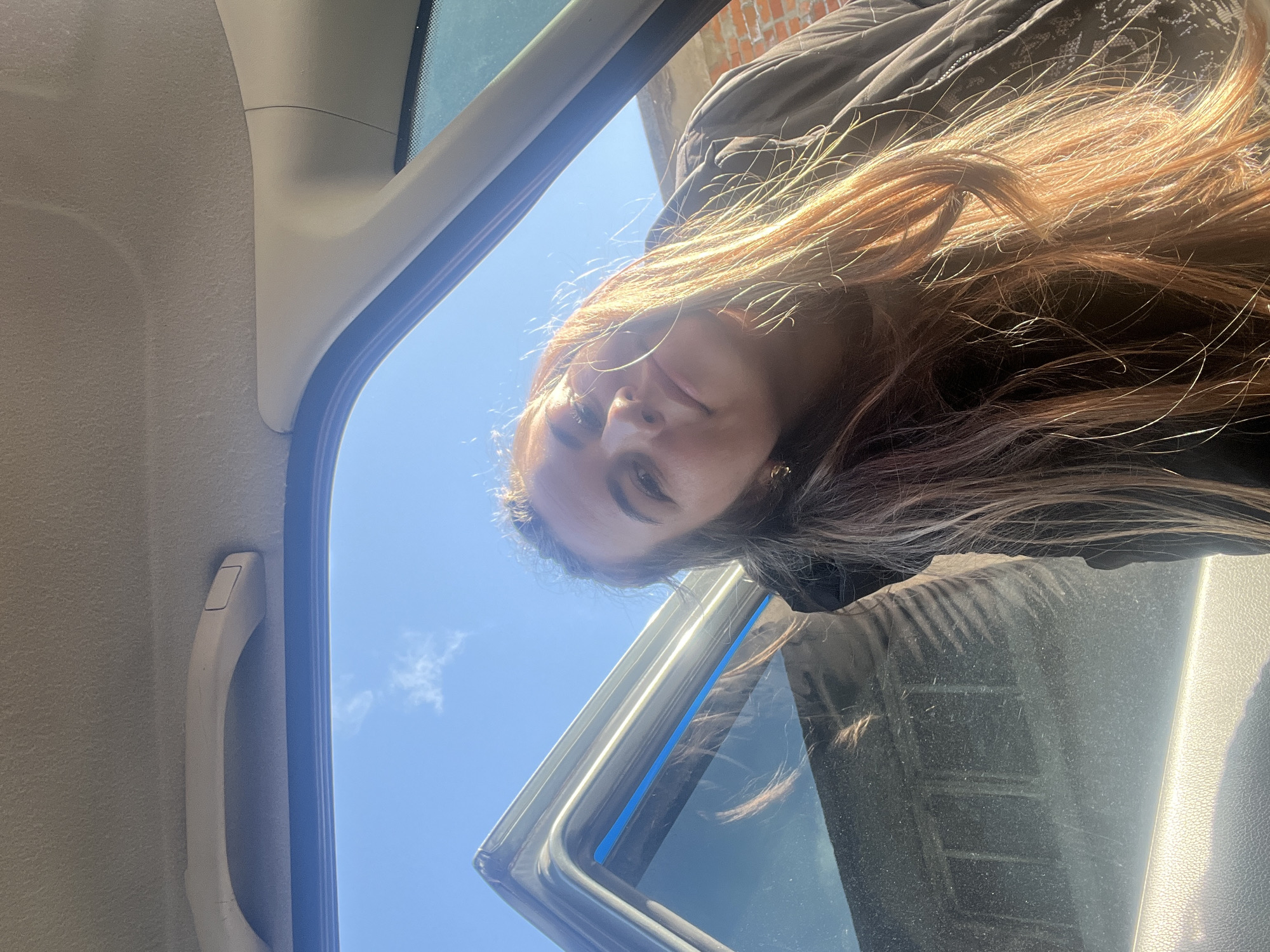 The image depicts a female subject captured from an interior perspective of a vehicle, looking into or leaning towards the vehicle. Bright daylight illuminates the scene under a clear blue sky with minimal cloud cover. The female has long, wavy, light brown hair, strongly backlit by the sun, and is wearing a dark, possibly quilted, jacket. She is looking directly towards the camera with a neutral expression.
Within the vehicle, a beige fabric headliner is visible at the top, along with a matching overhead grab handle. A light-colored plastic A-pillar trim and a portion of the front passenger window's dark rubber seal are also present. The exterior body of the vehicle, visible at the upper left, appears dark-colored with a distinct blue strip along the window edge. Through the window, in the background, a structure made of reddish-brown bricks is partially discernible, indicating an urban or residential environment. The location is within the city of Chaikovsky, Russia, characterized by this outdoor, urban setting visible from the vehicle.