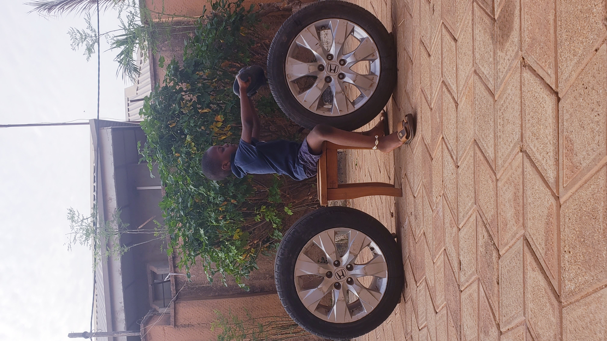 A young child, appearing to be a boy, is shown outdoors in Narayi, Nigeria, engaged in a playful activity. He is seated on a small, light brown wooden stool, dressed in a blue short-sleeved top and patterned shorts. An anklet adorned with cowrie shells is visible on his right ankle. The child is holding a dark, bowl-shaped object with both hands, raised above his head, mimicking a weightlifting or exercise pose.
Two car wheels with tires are prominent in the scene. They feature silver, multi-spoke rims, and the "Honda" logo is clearly visible on the hubcap of the wheel positioned behind the child. Another wheel is partially visible in the lower foreground. The ground beneath is paved with reddish-brown interlocking pavers arranged in a diagonal chevron pattern. Behind the child and the wheels, there is dense green foliage, suggesting a garden or yard environment. In the background, part of a building with a light-colored roof and walls can be seen, partially obscured by more sparse greenery, set against a bright, light-colored sky. The scene is illuminated by daylight, indicating a clear day.
