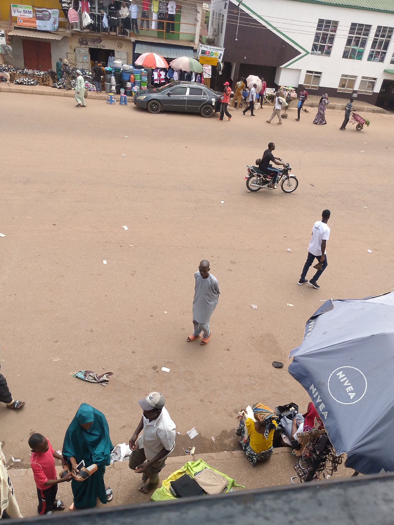 This photograph depicts a street scene in Bukuru, Nigeria. The ground is a dusty reddish-brown color, marked with scattered bits of paper and debris. In the upper left, several storefronts are visible, some displaying clothing and shoes for sale. A dark grey car is parked near these shops. Numerous people are walking or standing throughout the scene, some carrying items. A motorcyclist rides across the open area. At the bottom of the frame, there is a vendor seated near an open staircase, next to a Nivea-branded umbrella. A woman in a teal hijab, a child, and a man are looking at sandals.