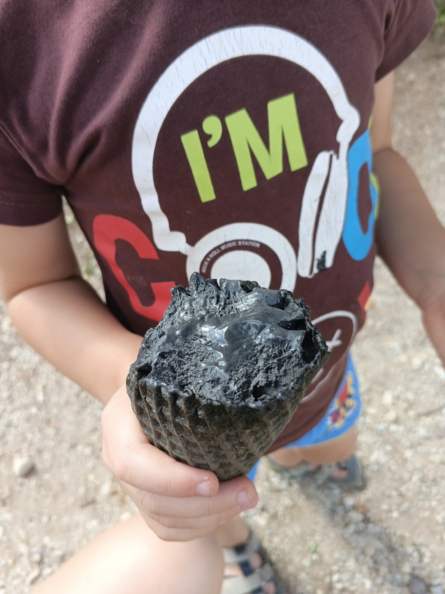 A person, possibly a child, is holding a piece of black, porous material that resembles a hardened dessert or a geological specimen. The material is dark gray to black with visible texture and some shiny, wet-looking patches. It is held in the person's left hand, with the fingers cupping the bottom and thumb supporting it. The person is wearing a maroon t-shirt with a white graphic that includes the text "I'M" in green and part of the word "GO." They are also wearing blue shorts with colorful patterns. The background consists of light-colored gravel and dirt, suggesting an outdoor, possibly rural or park-like setting. The image does not provide specific details to confirm the location in Medvedyovka, Russia.