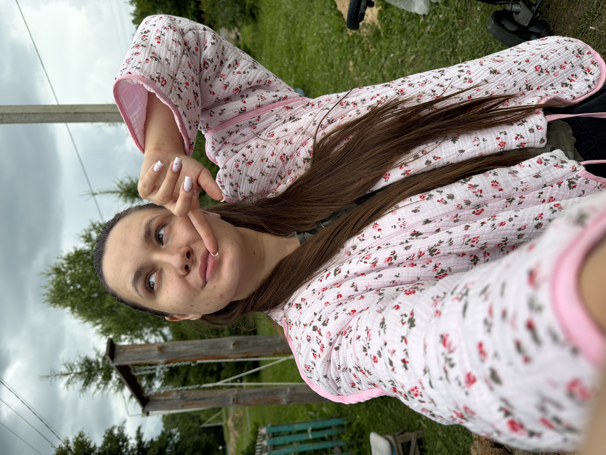 The media file is a selfie of a young woman in a pink floral robe taken outdoors in Krasny Mayak, Russia. The woman has long brown hair and is making a peace sign with her right hand near her face. She has light skin with some blemishes. The background shows a grassy area with trees and a wooden structure that appears to be part of a playground or swing set. The sky is overcast, suggesting it is daytime, possibly late afternoon. The overall mood is casual and relaxed, with the woman seemingly enjoying a moment outdoors. There is no visible text in the media file.