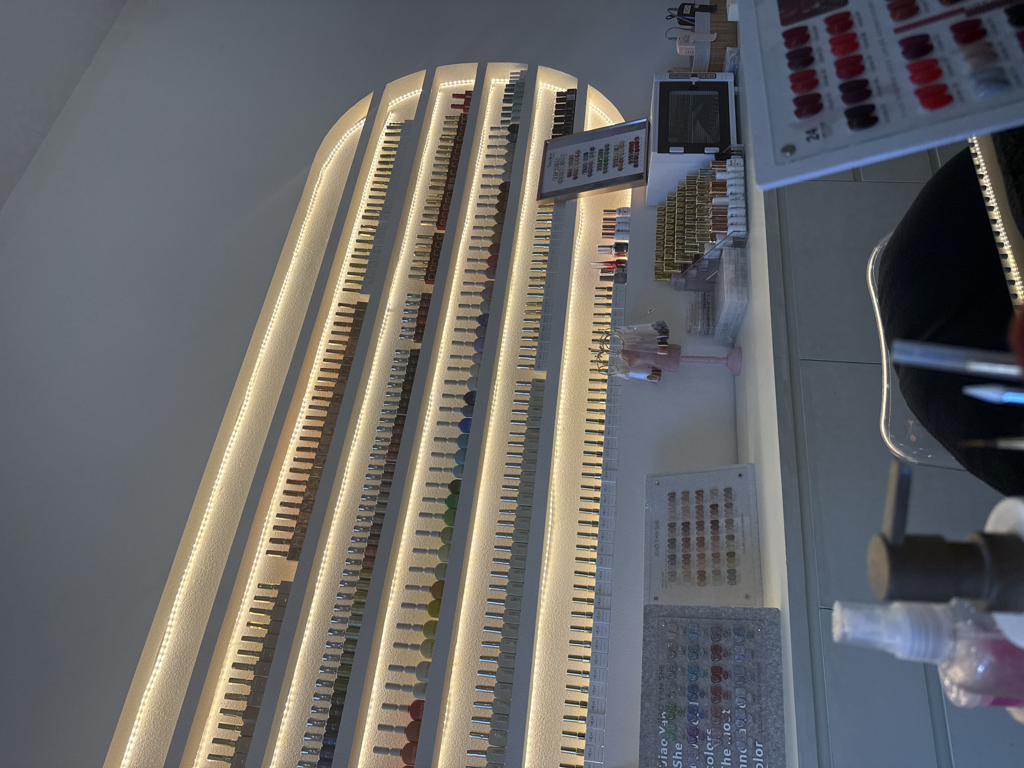 The image depicts an interior view of a nail salon, likely in North Kuta, Indonesia. The primary focus is a large, backlit shelf unit displaying numerous bottles of nail polish, organized by color and brand. The shelves are illuminated by warm, white LED strip lights, creating a soft glow that highlights the extensive collection. Below the shelves, a white countertop holds various salon supplies, including a color chart display, a small sterilizer or UV lamp, and several bottles of liquids, one of which is a pump dispenser. In the foreground, partially visible, is a person's lap and arm, suggesting they are seated at a station within the salon. The overall aesthetic is clean, modern, and geared towards showcasing a wide variety of nail polish options.