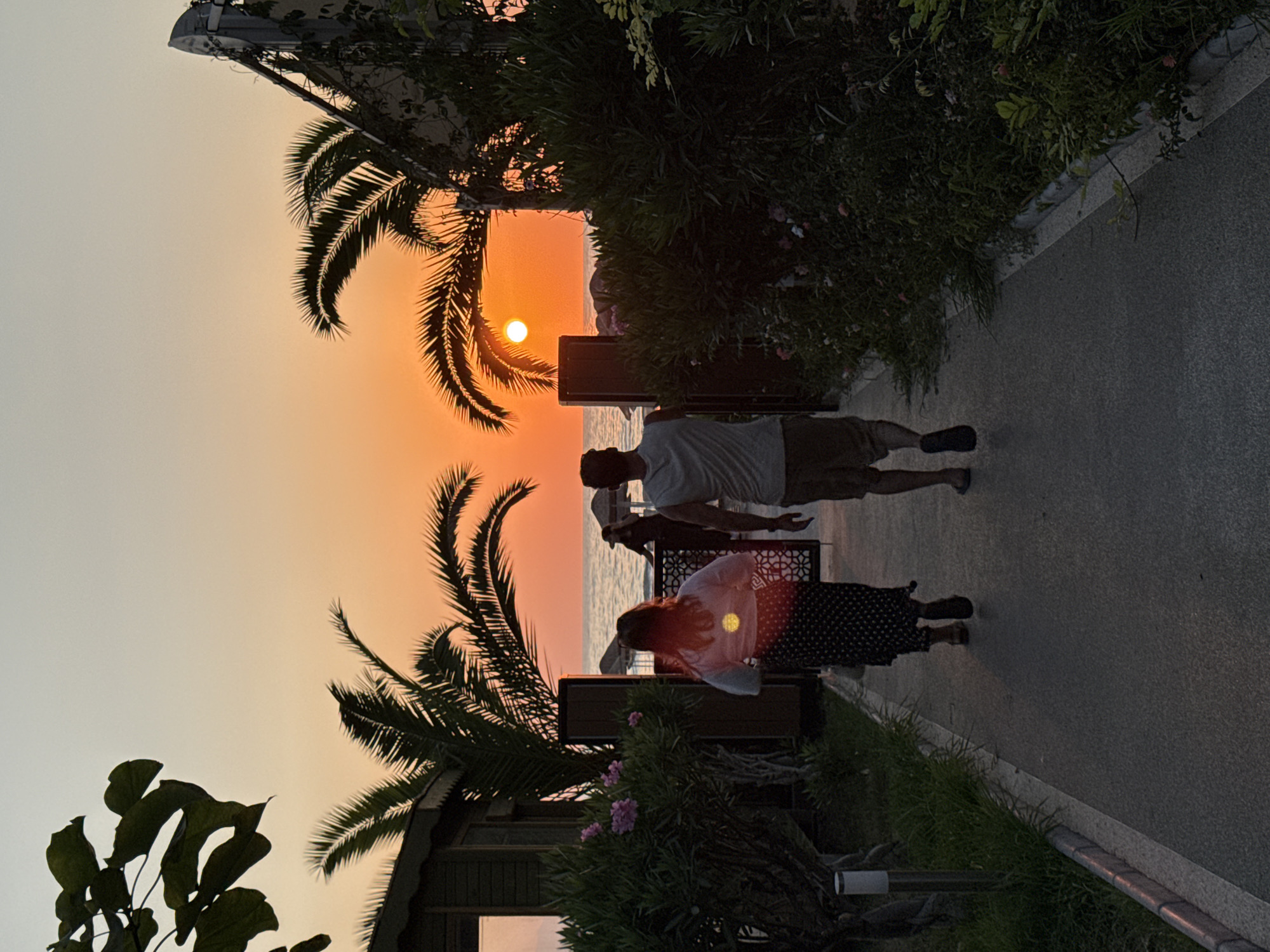 Two individuals are walking away from the camera on a paved walkway during sunset. The man on the right is wearing a grey tank top and khaki shorts, and the woman on the left is wearing a white top with a black and white polka dot skirt. They are heading towards the sea, which is visible in the background. The sky is a vibrant orange and yellow, characteristic of a sunset. Palm trees are on either side of the walkway, and there is lush green foliage and some pink flowers on the left. A small wooden building is partially visible on the left, and on the right, there's a structure with a curved roof and some vegetation. The scene appears to be a coastal resort area, possibly in Kuşadası, Turkey, given the palm trees and beach umbrellas in the distance, which are typical of such locations. The overall atmosphere is serene and picturesque.