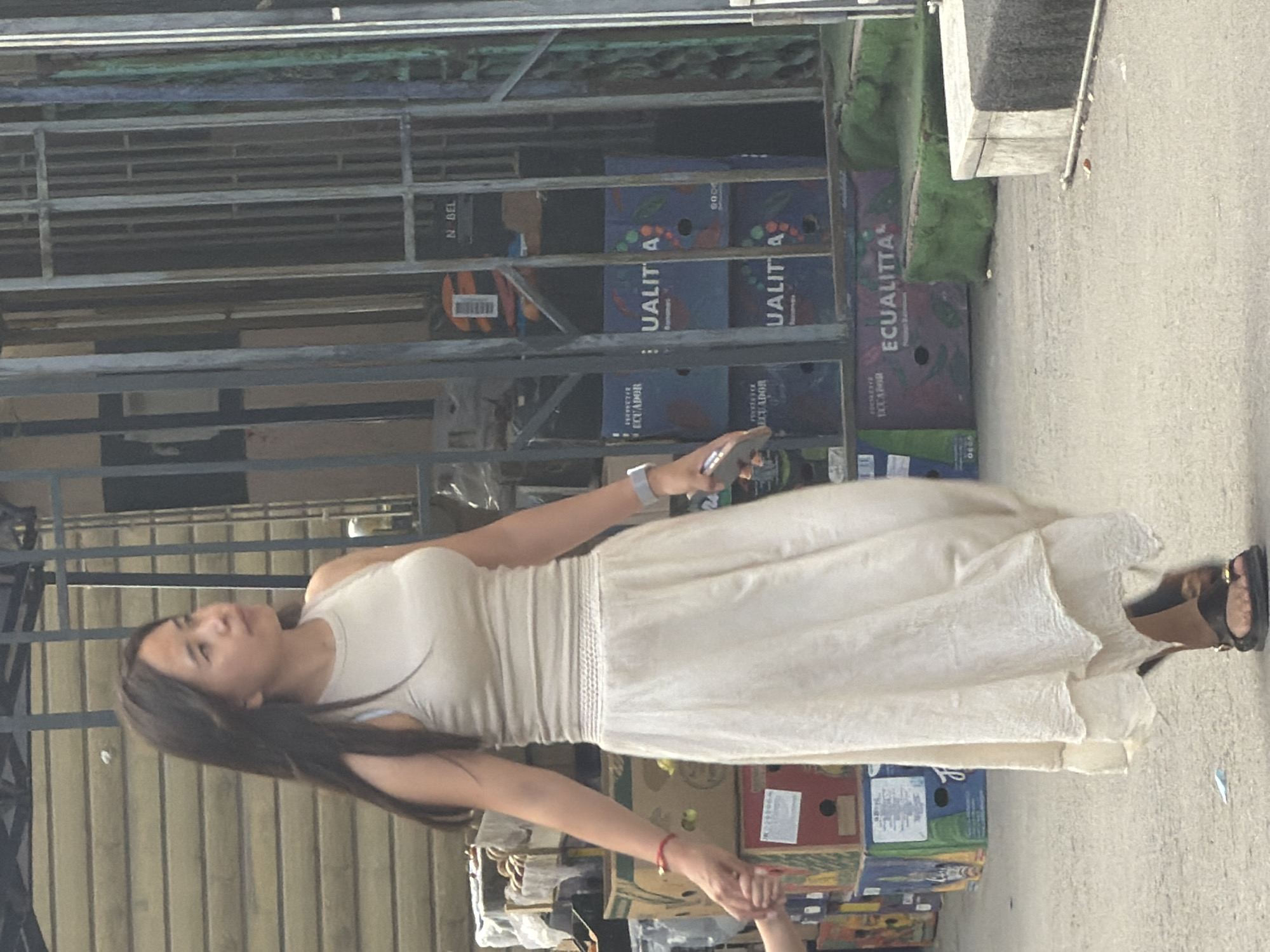 A young Asian woman with long, dark hair, wearing a light-colored tank top and a flowing, off-white midi skirt, stands in an outdoor setting, holding the hand of a child whose arm is visible on the left side of the frame. She is looking upwards and to the right, with a neutral expression. On her left wrist, she wears a red bracelet, and on her right wrist, a gray smartwatch. She is holding a mobile phone in her right hand.
Behind the woman, stacked boxes, some with labels like "ECUADOR" and "ECUALITTA" indicating bananas, are visible. A metal grate structure with vertical bars stands in the background, partially obscuring a light-colored wall. The ground is paved with a textured surface, and some green, moss-like material is present near the boxes. The overall scene appears to be a street or market area.