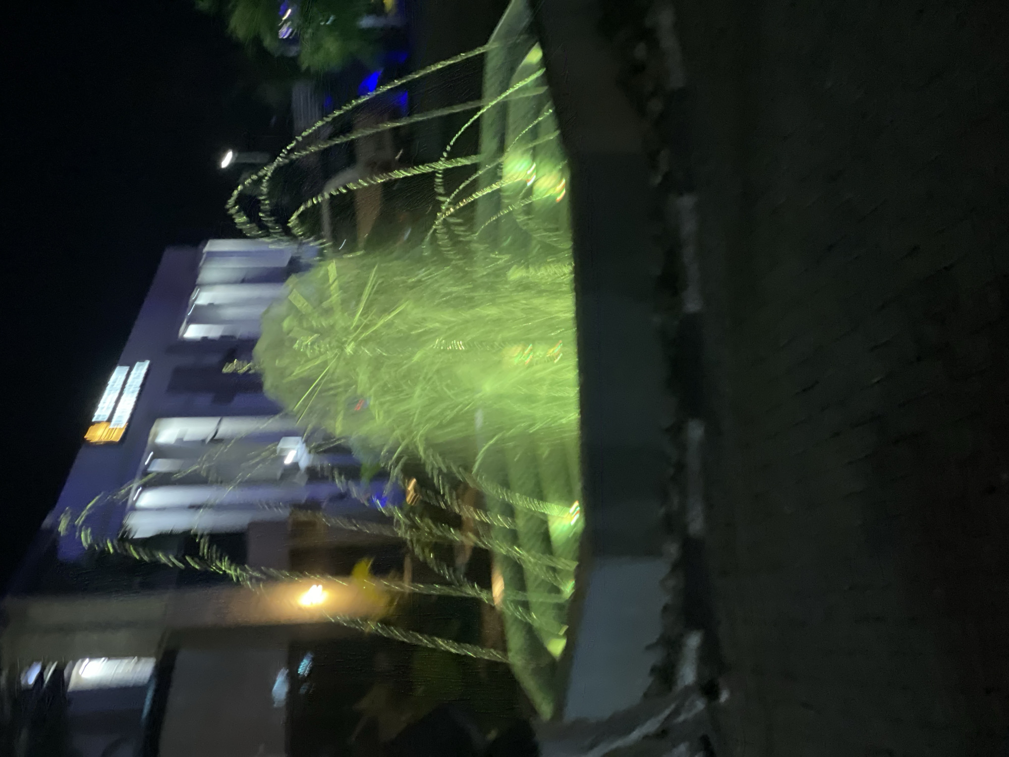 In Akwanga, Nigeria, at night, a fountain with green illumination is the primary subject. The fountain is centrally located and emits streams of water that are lit from below, creating a luminous green glow. The water arcs upwards and outwards, creating a dynamic visual effect. In the background, a modern building with large windows and vertical lighting is visible. The building appears to be multi-story and has signage at the top, though the text is not legible. The overall scene is set against a dark night sky, with some trees or foliage visible on the right side of the image. The ground in the foreground is paved with what appears to be brick or stone. The image has a slightly blurry quality, possibly due to low light conditions or motion. There are no people clearly visible in the image.