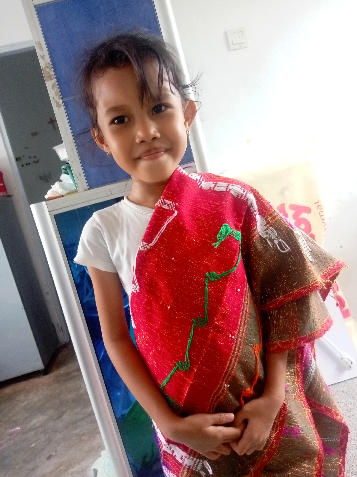 A young girl, appearing to be of Southeast Asian descent, stands in what appears to be an indoor setting. She is wearing a white t-shirt and a colorful traditional garment draped over her shoulders. The garment is predominantly red with intricate woven patterns in gold and green. Her hands are clasped in front of her. The background includes a blue and white tiled structure, a white wall with an electrical light switch, and a gray tiled floor. The overall lighting suggests daytime. The image is taken from a medium distance, focusing on the girl and her attire. The location is identified as Batam, Indonesia.