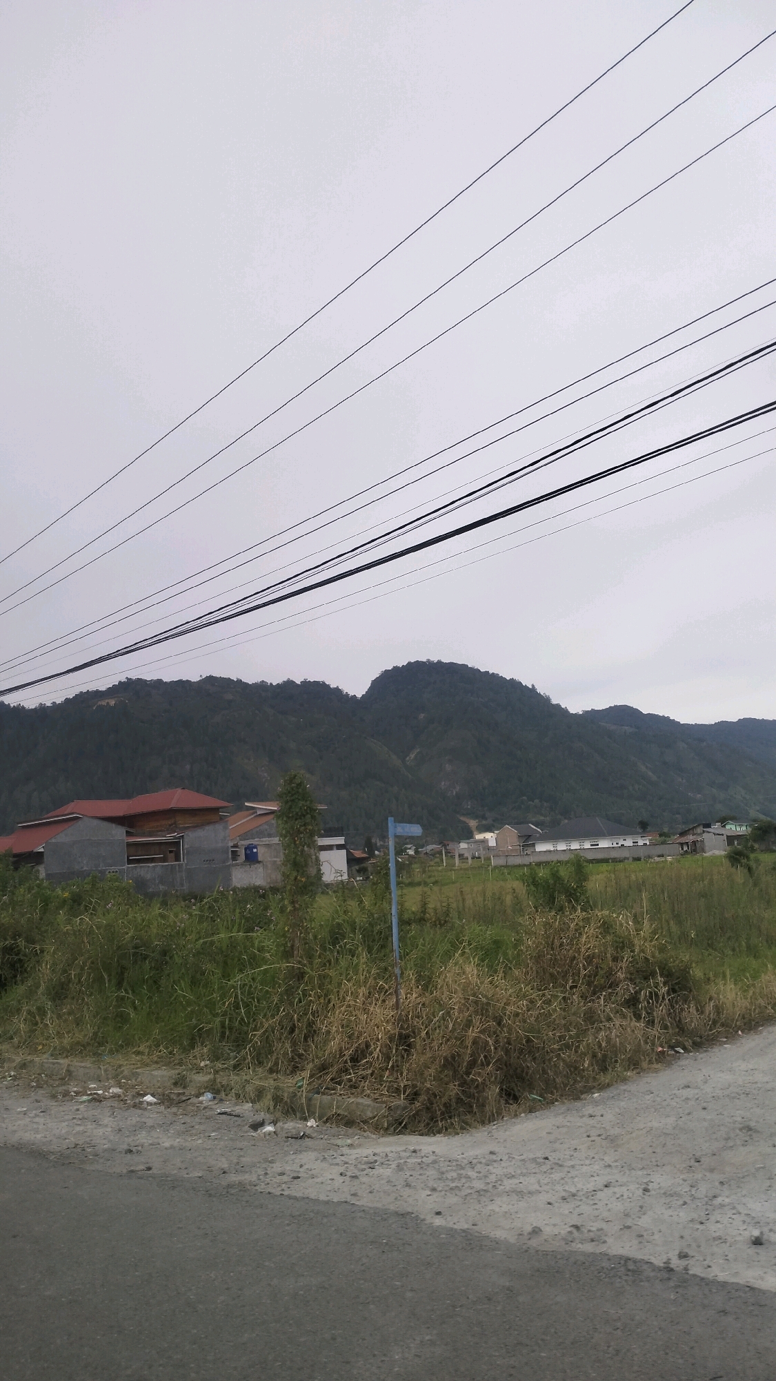 The image depicts a roadside view in Lotkala, Indonesia, with a prominent mountain range in the background under an overcast sky. Several power lines are strung across the upper portion of the frame. In the mid-ground, a cluster of houses with red tile roofs and gray walls are visible, alongside a small, dry, treed area. Further back, a residential area with several houses is spread across the lower slopes of the mountains. The foreground consists of a paved road and a gravelly shoulder, bordered by tall, dry grass and weeds, with some scattered litter. A blue directional signpost stands near the grassy area. The scene is quiet and appears to be a semi-rural residential area.