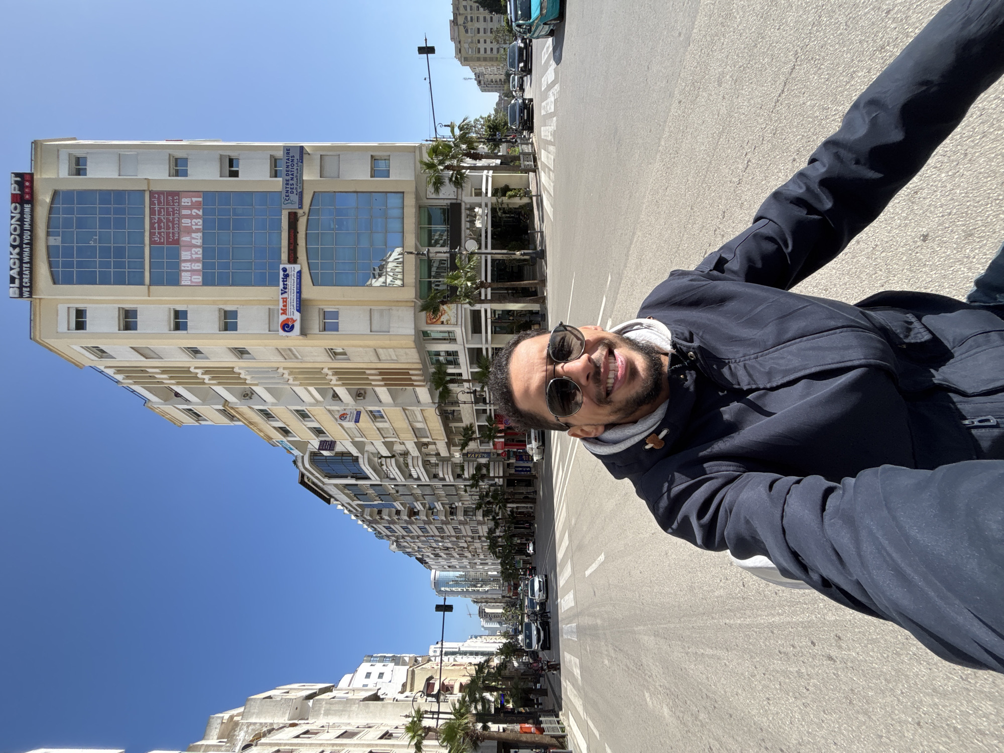 A male subject, approximately 30-40 years old, with short dark hair, a beard, and dark sunglasses, occupies the foreground, smiling at the camera in a selfie perspective. He wears a dark navy blue hooded jacket over a grey hoodie. The setting is a wide, multi-lane asphalt street with visible white dashed lane markings and a pedestrian crosswalk at an intersection.
The background features multi-story urban buildings under a clear blue sky. To the right, a prominent light-colored building, approximately 8-9 stories tall, features large blue reflective windows and several commercial signs. Visible signs include "BLACK OONO PT WE CREATE WHAT YOU IMAGINE" at the top, "Maxi Vertigo," and "CENTRE DENTAIRE DES NATIONS." Another sign displays "BUREAUX A LOUER" (Offices for Rent) with a telephone number (06 13 44 13 2 1) in Arabic and French. Palm trees line the sidewalk in front of this building. The left side of the street is lined with similar multi-story buildings extending into the distance.
Moderate vehicle traffic is present on the street, including several sedans, a minivan, an SUV, and a teal-colored compact car. A black traffic light signal is visible above the intersection.
The location is identified as Place des Nations Unies, Tangier (Tanger), Morocco, specifically near the building housing "CENTRE DENTAIRE DES NATIONS" and "BLACK OONO PT." The perspective likely faces down Avenue du Prince Héritier.