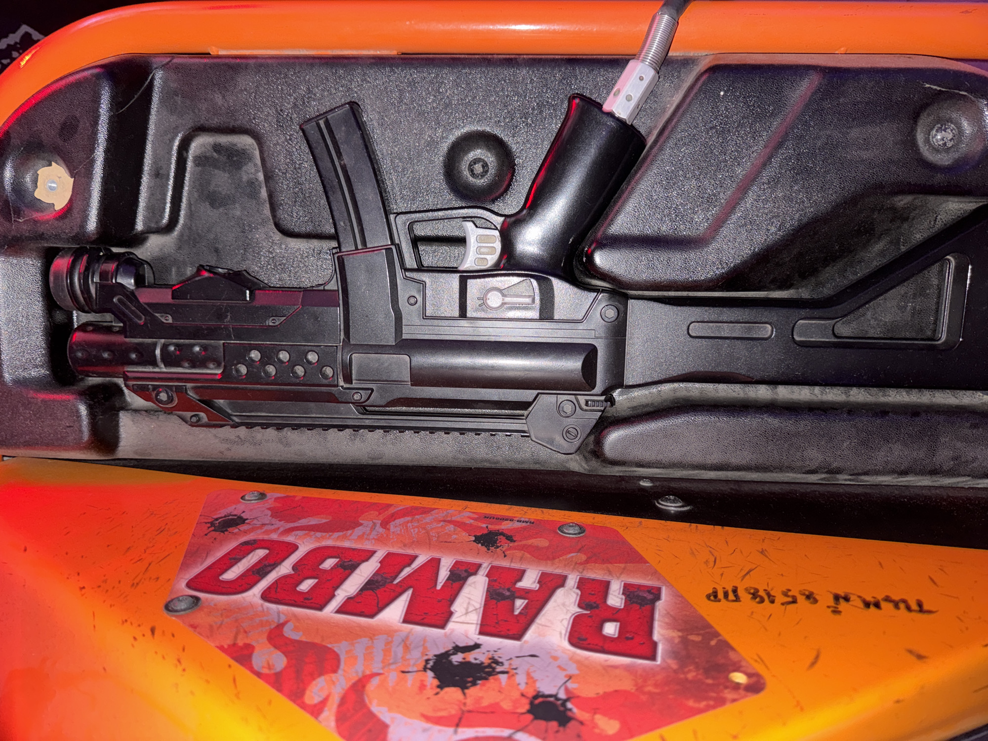 The image shows a close-up, overhead view of a toy gun placed inside an orange and black container. The toy gun is primarily black with some silver accents. It has a detailed design, resembling a realistic firearm with a magazine, trigger, and barrel. The gun is positioned horizontally within the container. The container itself appears to be made of plastic, with textured black surfaces and a bright orange rim visible at the top. The background is dark, suggesting an indoor setting.
The lower part of the image reveals a bright orange surface with a graphic design. This design features the word "RAMBO" in large, bold red letters with a distressed effect. There are also stylized flames and black splatters, possibly representing bullet holes or ink blots, surrounding the text. Tiny, faint scratches are visible on the orange surface. There is also some small, handwritten text in black on the orange surface, which appears to be "TUMRI 8518 NP" written in a mirrored, backward direction.
There are no people visible in the image. The scene is static, with the toy gun simply placed within the container. The lighting appears to be artificial, with red light reflecting on the toy gun and parts of the container, suggesting a possible light source from below or the side. The overall impression is that of a prop or toy, possibly part of a game or collection, with the "RAMBO" branding strongly suggesting a connection to the movie franchise. The location context is Novaya Usman, Russia, but this cannot be visually inferred from the image itself.