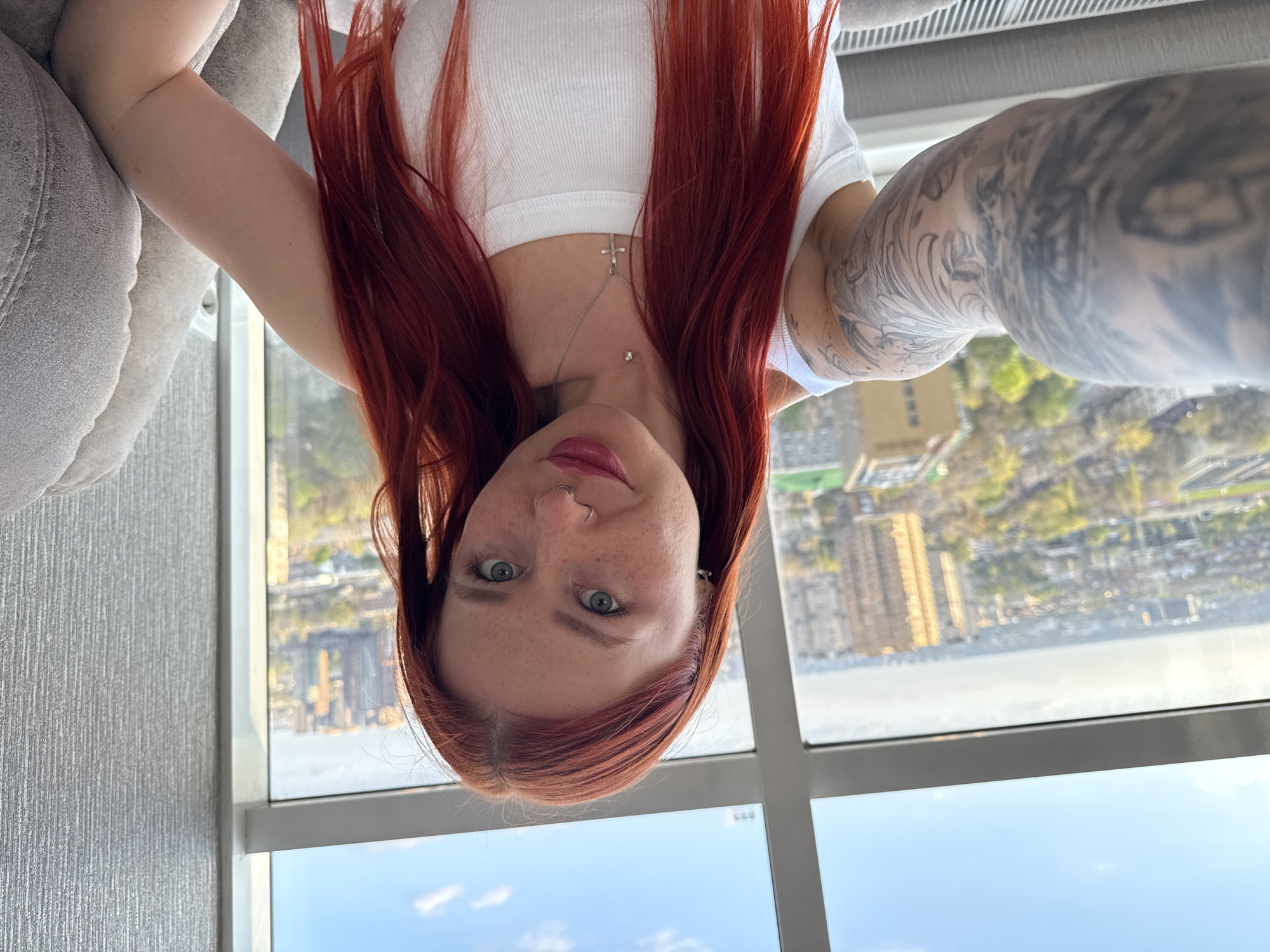 A young woman with red hair and freckles takes a selfie indoors, by a large window overlooking the cityscape of Rostov-on-Don, Russia. She wears a white tank top and a necklace with a cross. Her right arm is visibly tattooed. The scene shows part of a sofa and a clear view of high-rise buildings in the distance, reflecting in the windows on a bright day.
