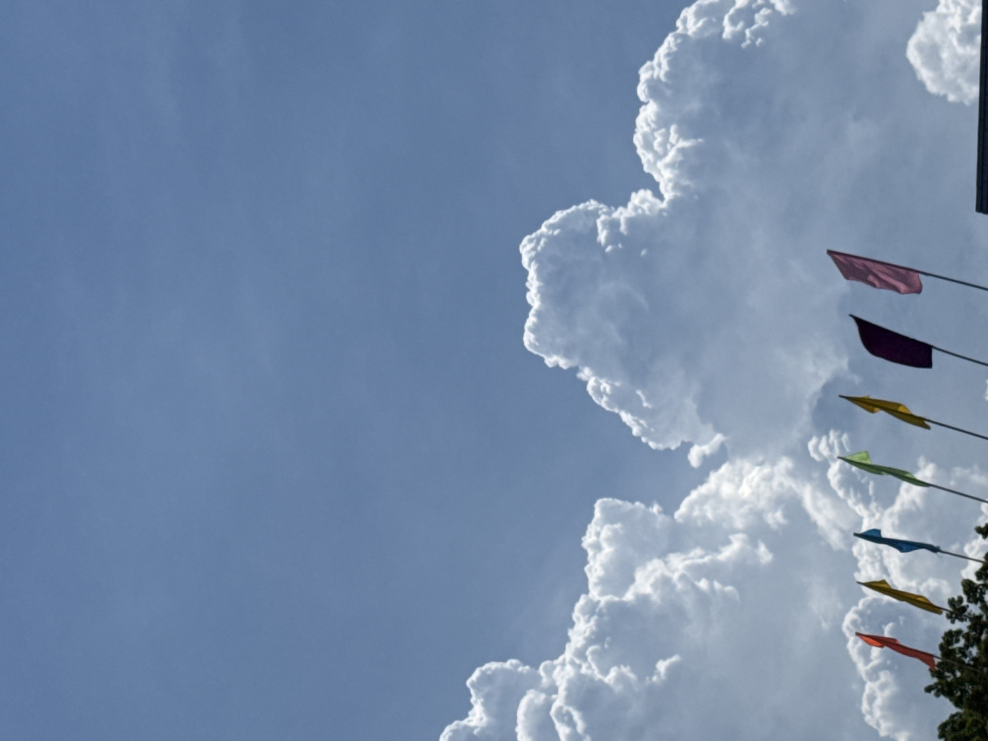 This media file features a bright, partly cloudy sky with a few colorful flags. The clouds are predominantly white and puffy, suggesting fair weather with some potential for accumulation, possibly cumulus clouds developing. The sky is a clear, bright blue, indicating it's likely daytime.
On the right side of the frame, a series of small, rectangular flags are visible, each attached to a thin pole. The flags display a variety of colors, including a dark red, a deep purple, a bright yellow, a lime green, a dark blue, another yellow, and a vibrant red at the bottom. These flags are arranged diagonally, ascending from the lower right corner towards the upper right. They appear to be blowing in a gentle breeze, their fabric rippling.
In the extreme lower right corner, the dark green foliage of a tree is partially visible, suggesting an outdoor setting. The overall impression is one of a pleasant, possibly festive day, with the bright colors of the flags contrasting with the vastness of the sky and the dramatic cloud formations. There is no visible text in the image. The scene evokes a sense of openness and mild weather, suitable for an outdoor gathering or celebration. The location context provided, Bac Nha Trang Ward, Vietnam, might suggest this could be a coastal or tropical environment, which would be consistent with the bright sky and puffy clouds.
