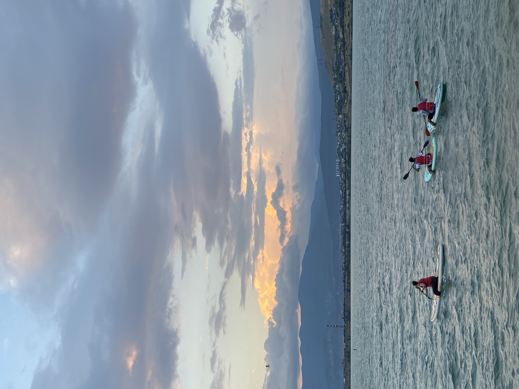 The image captures a serene evening scene on the Tbilisi Sea, a large reservoir on the outskirts of Tbilisi, Georgia. Three individuals are seen paddleboarding on the calm, rippling water. Each person is wearing a red life vest and is actively paddling their board. In the background, a range of mountains forms a scenic backdrop, with a distant cityscape nestled along the foothills. The sky above is a dramatic canvas of clouds, with patches of sunlight illuminating the horizon, suggesting either sunrise or sunset. The overall atmosphere is one of peaceful recreation against a natural and urban landscape.