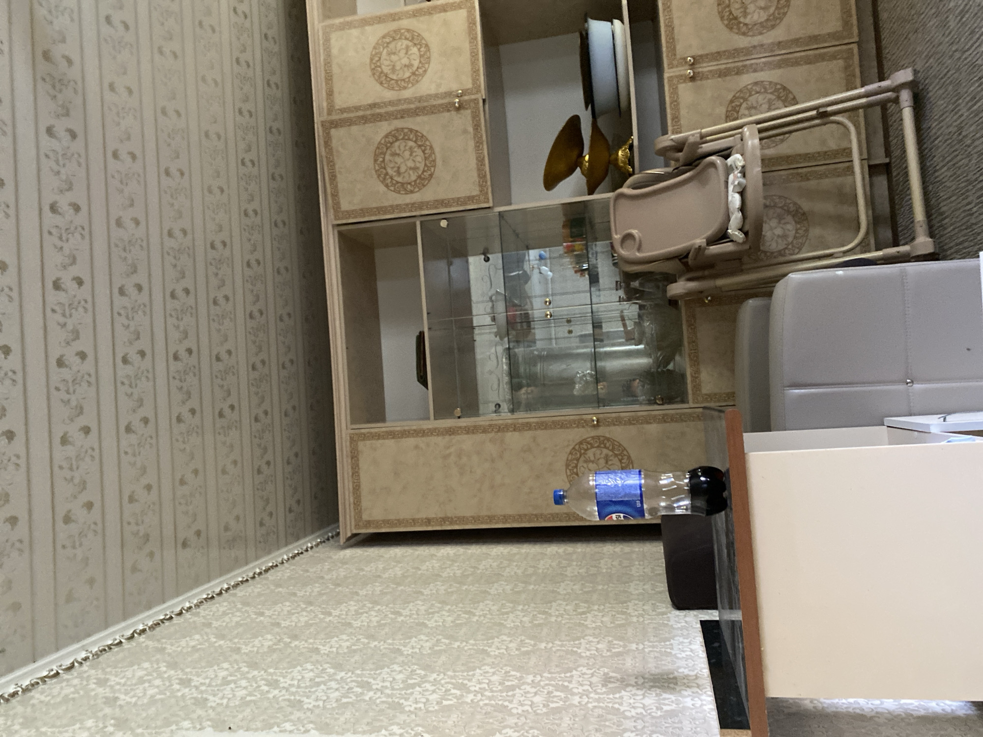 The image shows an indoor scene, likely a living room or dining area. A large, ornate cabinet with glass doors and decorative panels dominates the right side of the frame. On a counter in front of the cabinet, a partially filled bottle of Pepsi is visible. To the left of the counter, a laptop is present. In the foreground, a beige baby high chair is folded and leaning against the cabinet. The walls are covered in patterned wallpaper, and the ceiling features a decorative embossed pattern. The flooring appears to be carpeted. The overall impression is of a domestic setting in Миң-Булак, Kyrgyzstan.