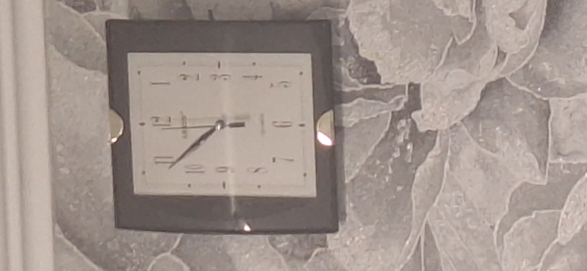A square wall clock is mounted on a textured wall. The clock face is white with black numerals and hands. The hour hand is pointing to approximately 10, and the minute hand is pointing to approximately 3. The clock has a dark gray frame. A bright light is reflecting off the bottom edge of the clock frame. The wall has a light gray, textured pattern resembling floral or abstract shapes. There is a white molding at the top of the wall. The image is taken from a slightly low angle. The location of this image is not provided.