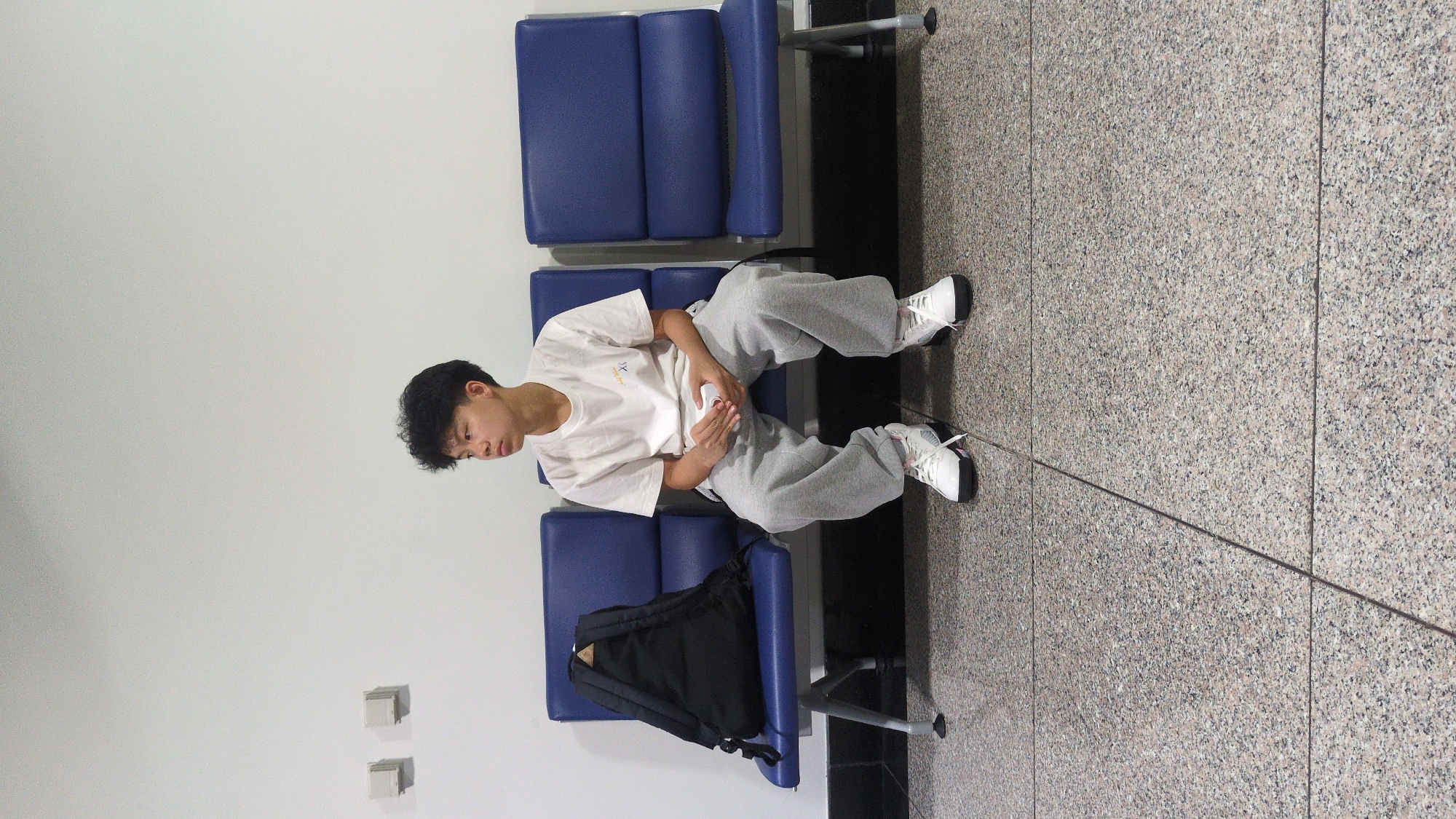 A young Asian male is seated on blue public waiting area chairs in an indoor setting in Thuận An, Vietnam. He is dressed casually in a white short-sleeved t-shirt with a light brown or golden stylized logo (possibly "XL") on the chest, light grey sweatpants, and white athletic shoes with black accents. He holds a white object, likely a smartphone or small device, in both hands and looks down at it, appearing engrossed or patiently waiting.
A black backpack rests on the seat next to him. The seating is composed of multiple connected blue vinyl chairs, commonly found in public transport hubs like airports or bus stations, or waiting rooms. The floor is made of light-colored, speckled granite-like tiles, and a thin, dark cable or line extends across it. The walls are plain white. The overall scene suggests a moment of quiet waiting or relaxation in a public facility.