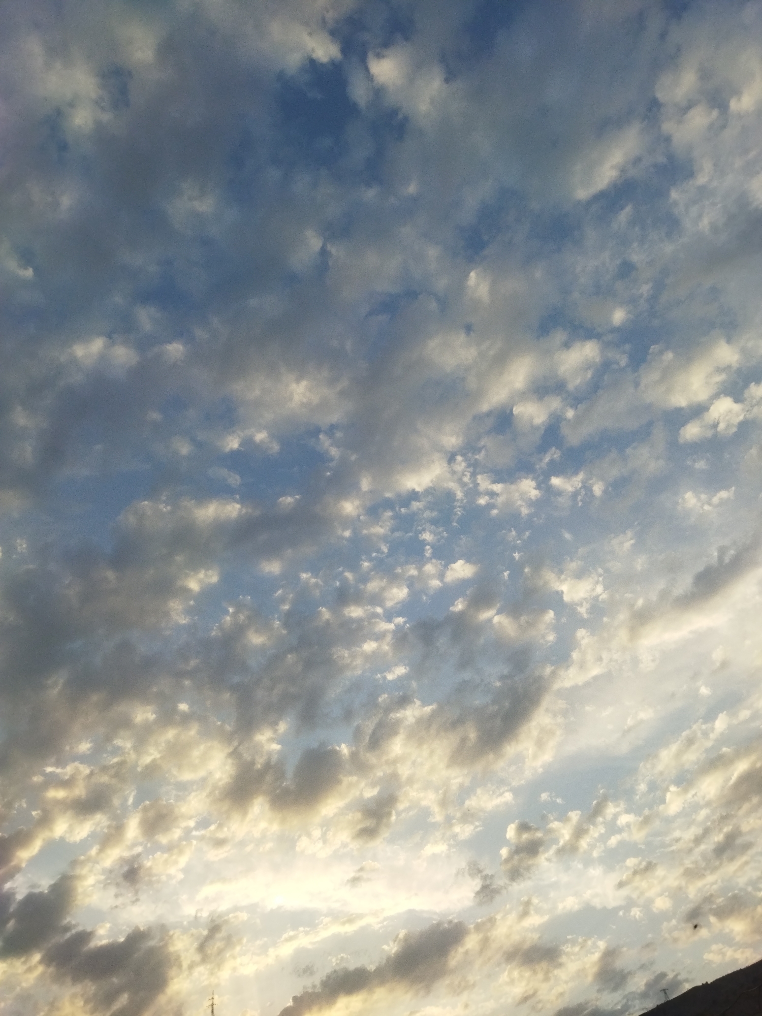The image shows a sky filled with clouds, with patches of blue sky visible between them. The clouds are varied in shape and density, with some appearing bright and illuminated by sunlight, while others are darker and more shadowed. The lighting suggests it might be during sunrise or sunset, as some clouds are tinged with yellow and gold. The lower portion of the image shows a sliver of land and a faint silhouette of a transmission tower, indicating a likely outdoor scene in a somewhat rural or semi-urban environment. There are no people or overt human activity visible in the image, only the natural elements of the sky and a hint of infrastructure. The scene appears to be an unobstructed view of the sky, possibly captured from a higher vantage point overlooking a landscape.