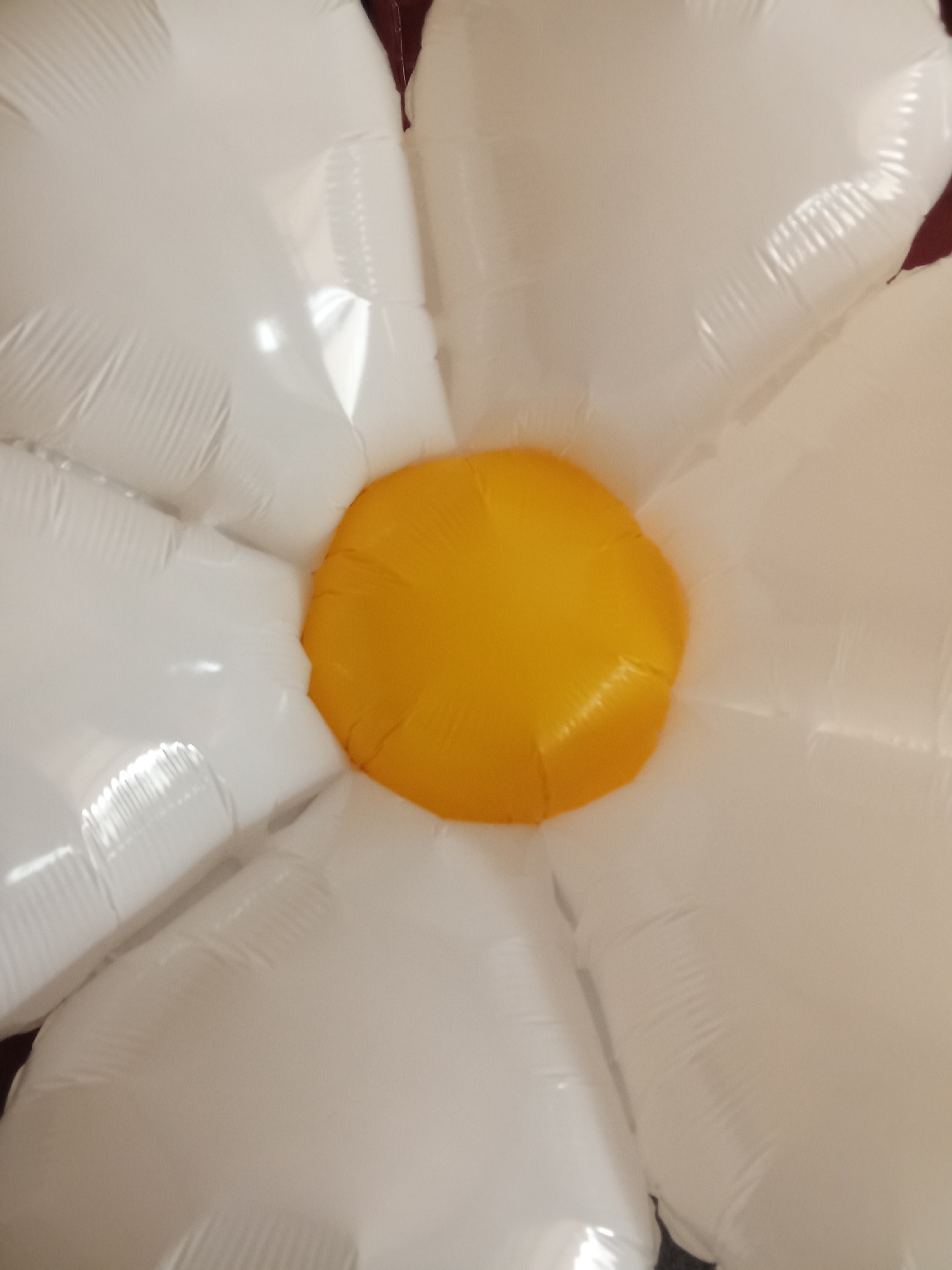 A close-up, overhead shot shows a large, inflatable daisy-shaped balloon. The center of the daisy is a bright yellow, round area, likely made of vinyl or a similar material. Radiating outwards from the yellow center are several large, white, petal-shaped sections, also appearing to be made of inflated material. The petals have a slightly creased texture, indicating they are filled with air. The background is dark and out of focus, suggesting the balloon is indoors, possibly in a residential setting in Tanah Mas, Indonesia. There are no people visible in the image.
