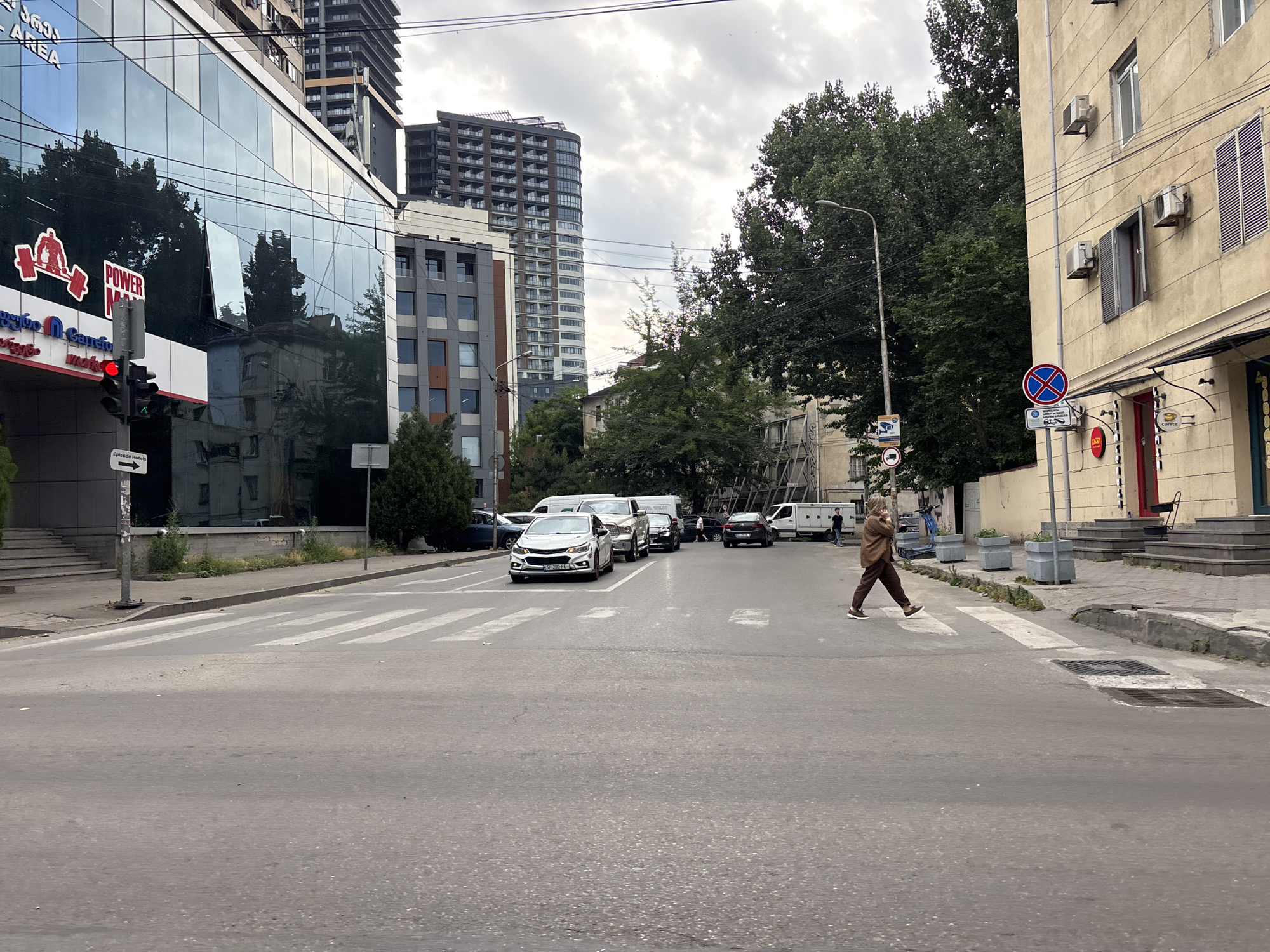 The image depicts a street scene in Tbilisi, Georgia, under an overcast sky. In the foreground, a pedestrian is crossing a crosswalk from right to left. The person is wearing a brown jacket and pants, and white sneakers. Behind the pedestrian, several cars are stopped in traffic, including a white SUV and a silver sedan. A white van is parked on the right side of the road.
The left side of the image features a modern building with a glass facade, reflecting the trees and sky. A red traffic light is visible, along with a sign indicating "Episode Hotels". Further down the street, a tall, contemporary building stands out against the cloudy sky, surrounded by trees.
On the right side of the street, a yellowish building with visible air conditioning units and shutters is present. Street signs, including a "no stopping" sign, are also visible. The overall impression is a busy urban environment with mixed architectural styles, common in Tbilisi. The road surface is asphalt, and the crosswalk markings are clearly visible.